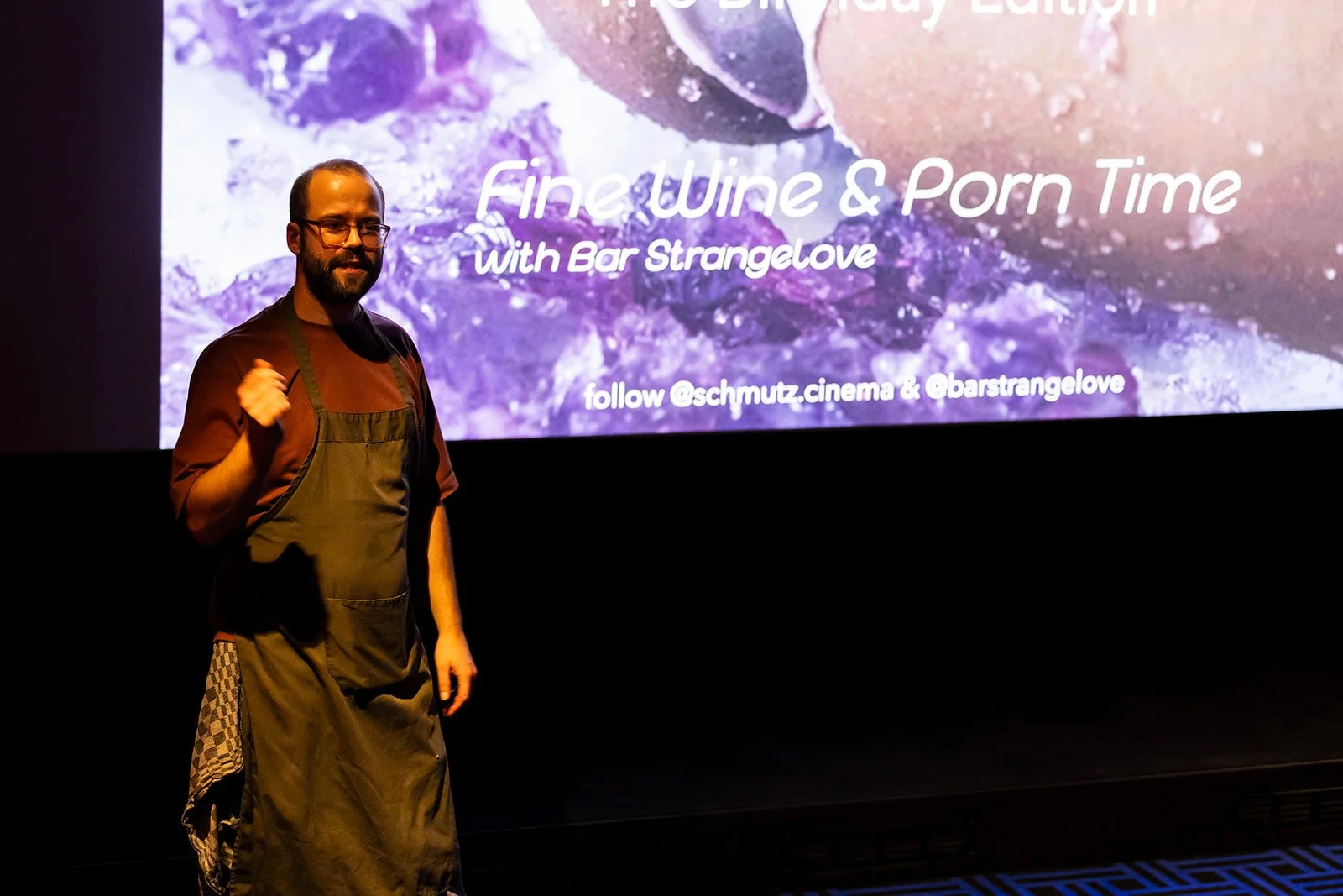 A man wearing glasses, a maroon shirt, and a dark apron stands in front of a large projection screen displaying text about wine and porn. The background of the screen features purple and pink hues.