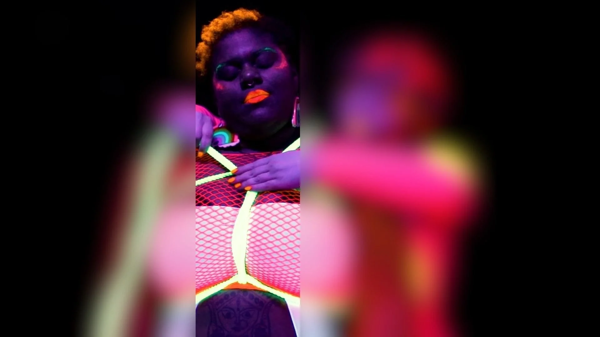 A person with dark skin has face painted in vibrant colors, with orange lips and purple face paint, and is wearing neon-colored accessories. They are wearing a brightly colored, mesh-like top with neon accents and have their hand placed on their chest.