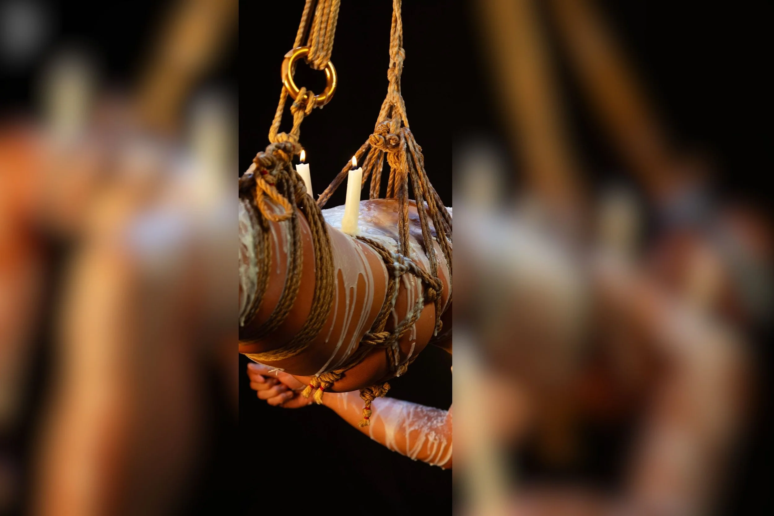 A person bound and suspended by thick shibari ropes, holding a candle and candles are placed on top, with a dark background.