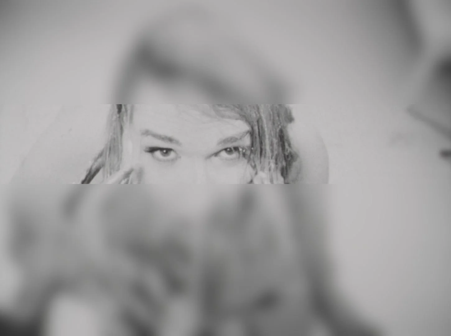 Black-and-white photo of a woman with dreadlocks and striking eyes, peering through a rectangular gap in a blurred background.