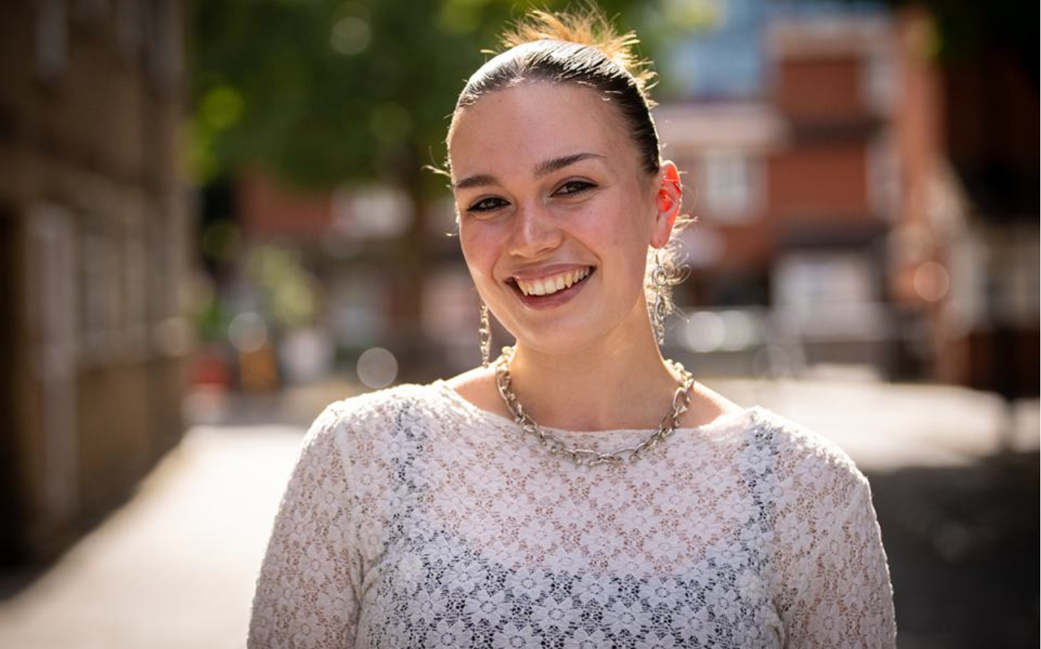 A young woman smiling outdoors on a sunny day, wearing a white lace top and jewelry, with a blurred urban background.