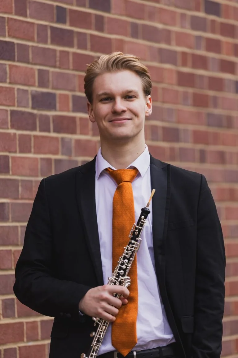 A young man in a black suit and white shirt holding a clarinet, standing outdoors in front of a brick wall, smiling.