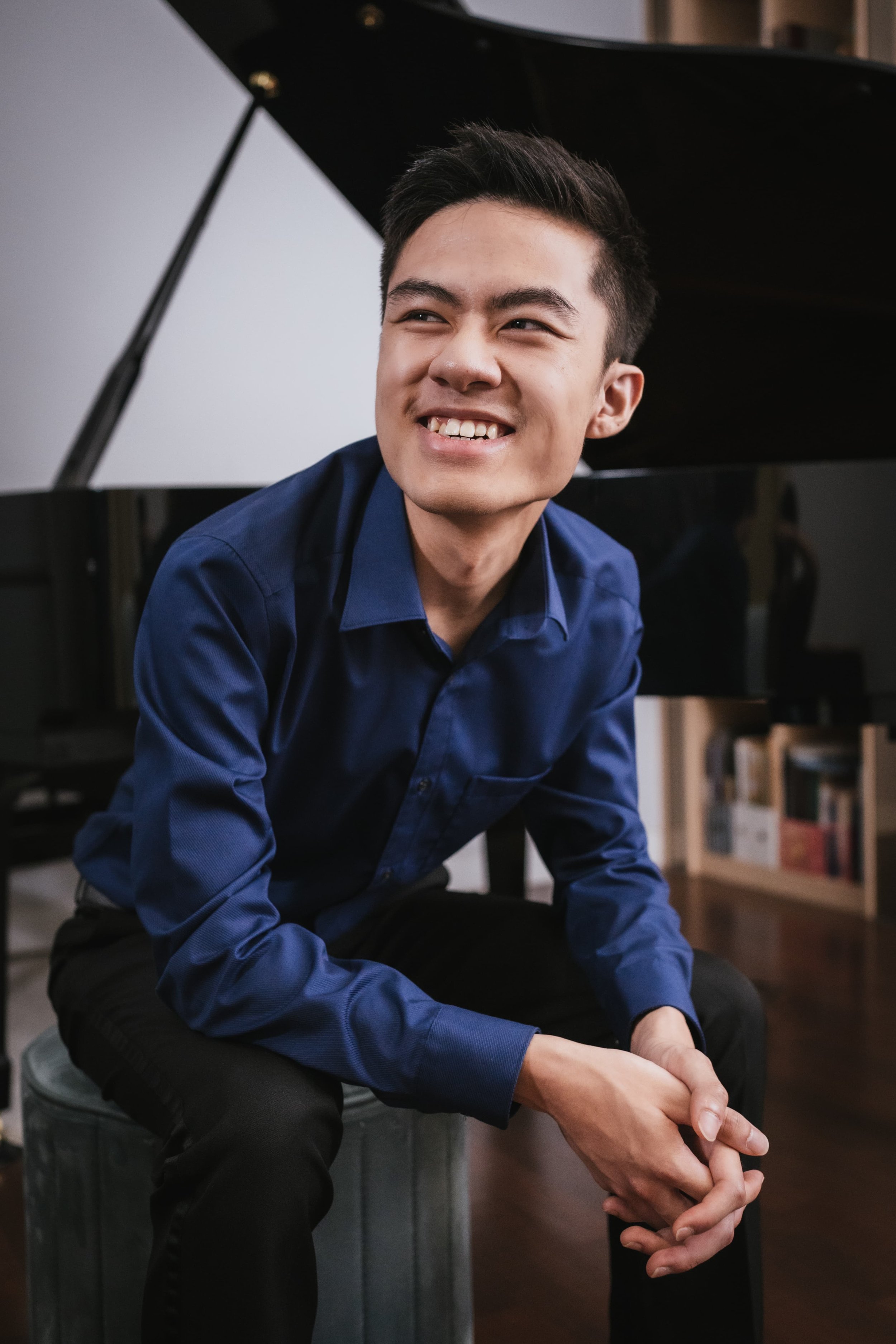 A young man smiling slightly, dressed in a dark blue shirt, sitting in front of a black grand piano, with a background of bookshelves.