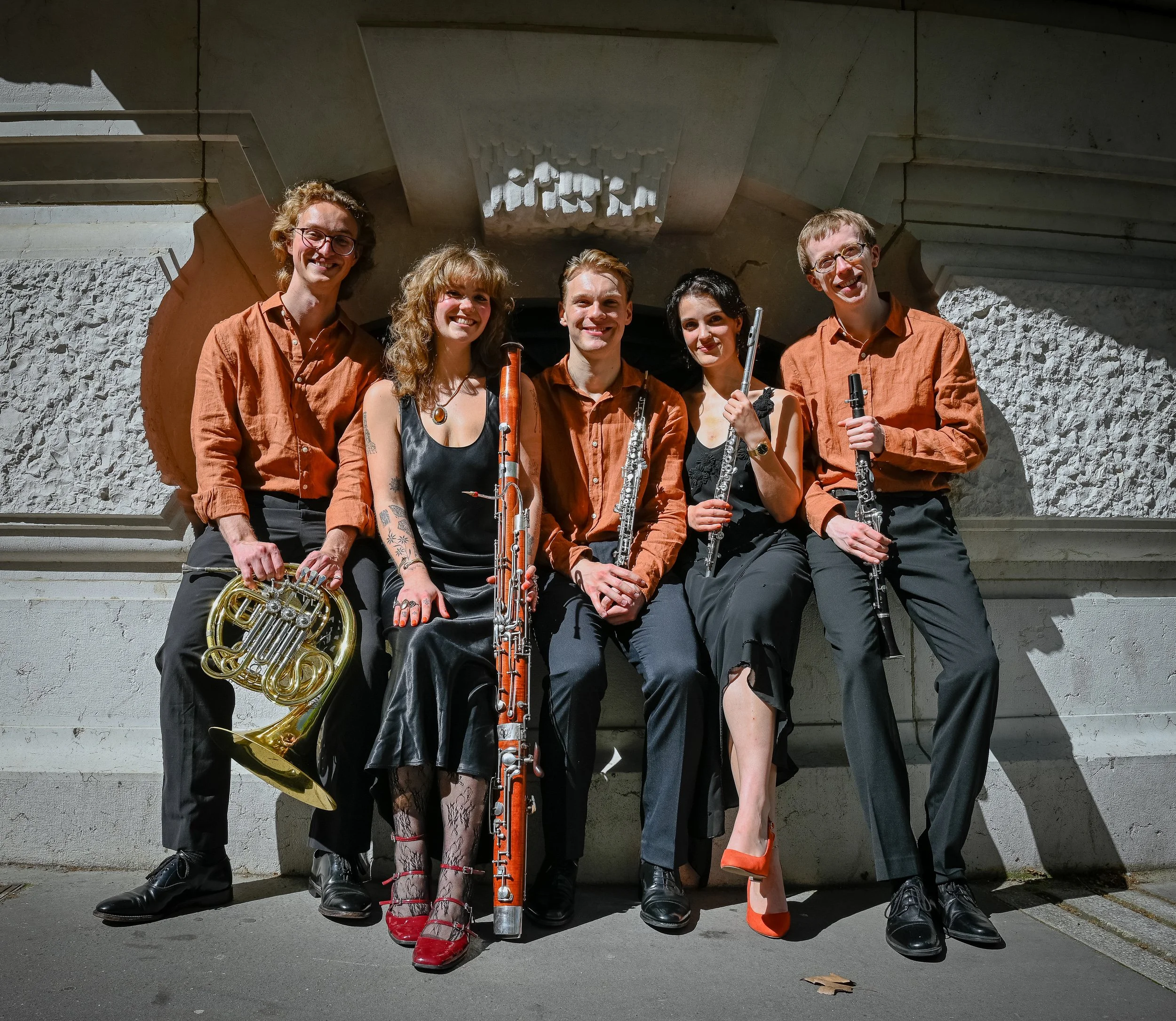 Group of five musicians standing outdoors against a concrete wall, holding musical instruments including a French horn, a flute, an oboe, a clarinet, and a bassoon, all dressed in matching orange shirts.