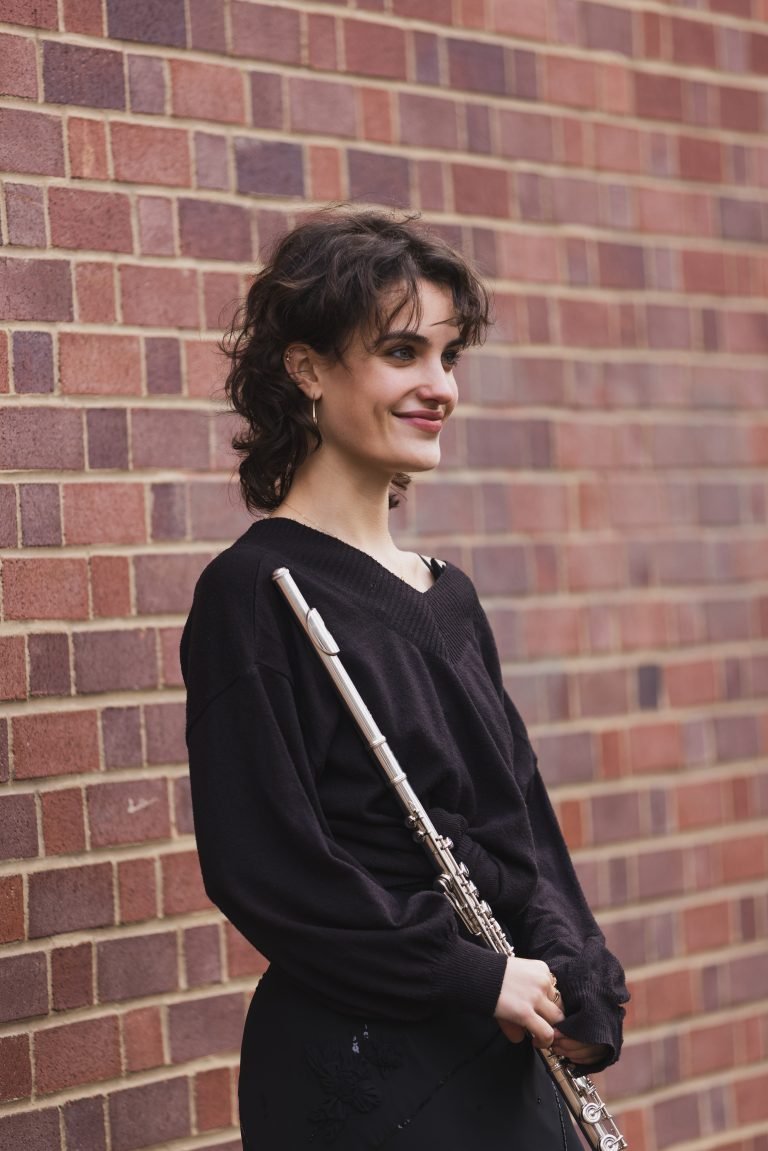 Young woman with short curly brown hair, wearing a black sweater and earrings, standing against a brick wall, holding a flute.
