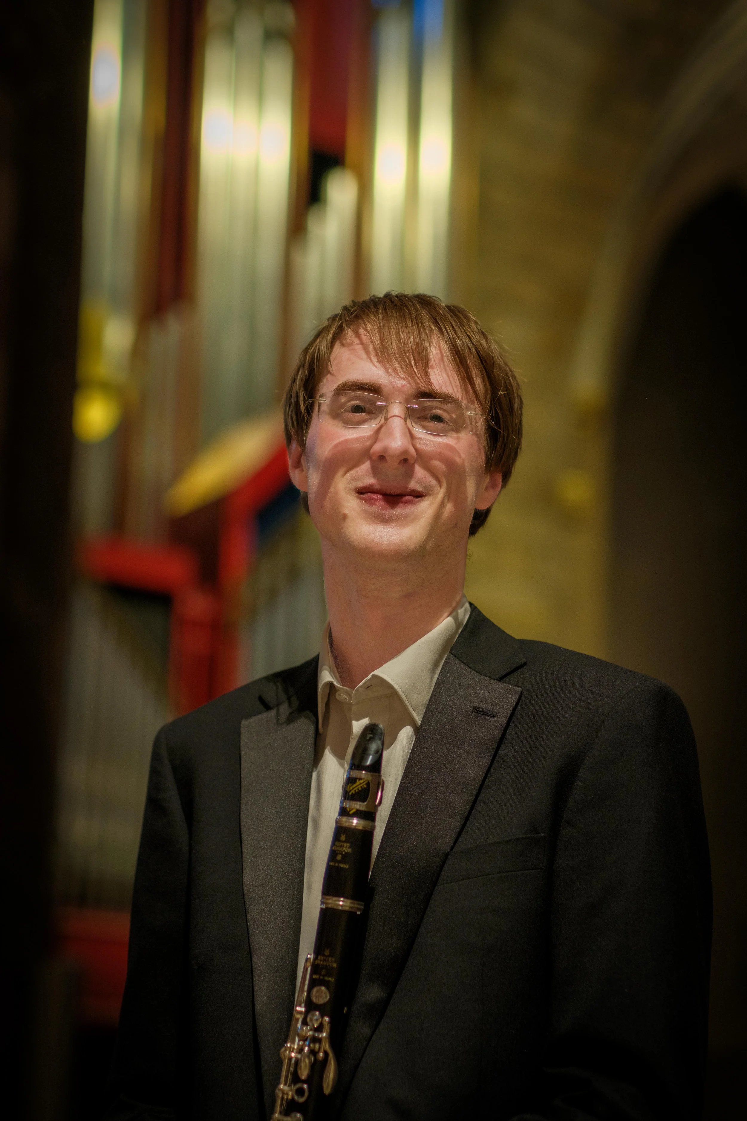 A man in a tuxedo holding a clarinet, standing inside a hallway with colorful stained glass windows.