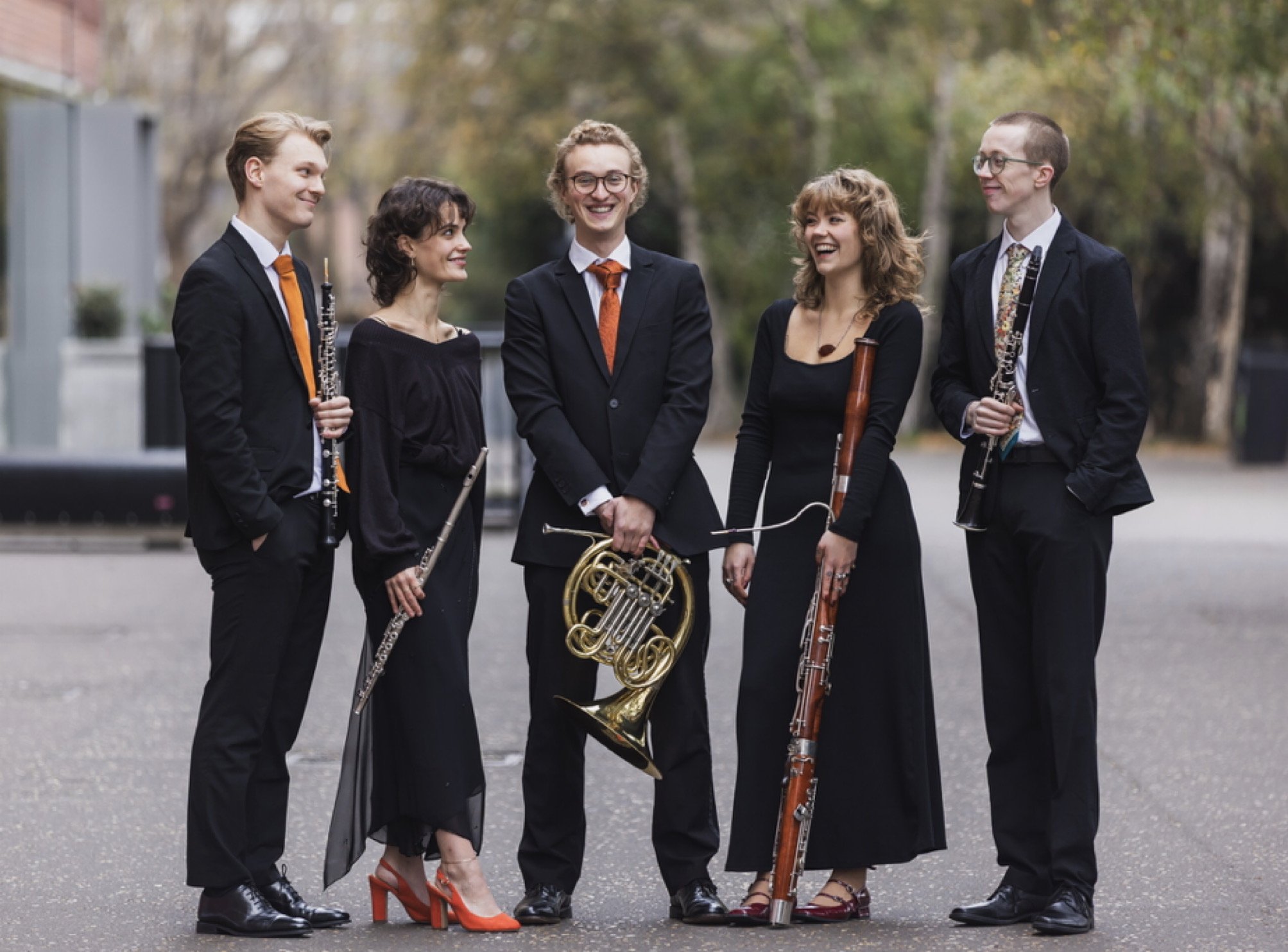 Five musicians, three men and two women, standing outdoors on a paved area with trees in the background. They are dressed in formal black attire and holding musical instruments including a clarinet, French horn, bassoon, and another clarinet, and are smiling and engaging in conversation.