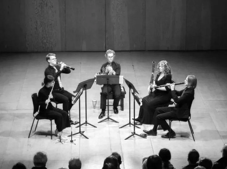 Five musicians playing woodwind instruments on stage during a performance, with audience visible in the foreground.