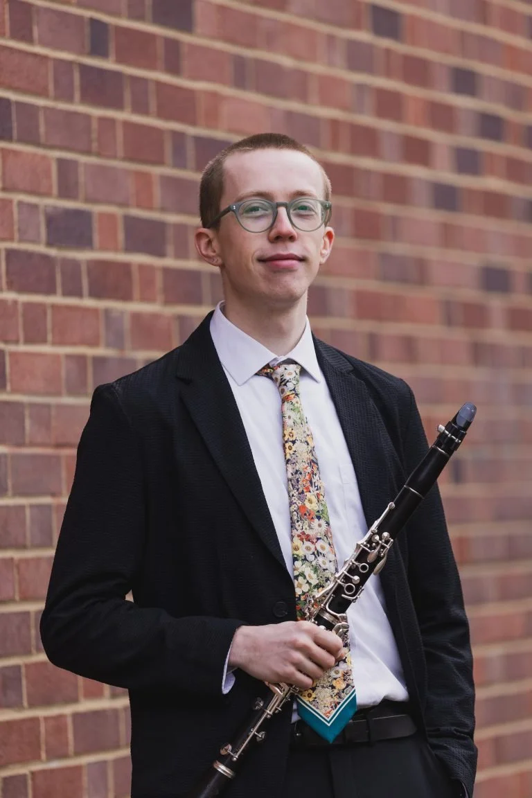 Young man in glasses holding a clarinet, standing outdoors against a brick wall, wearing a white shirt, black blazer, and a floral tie.