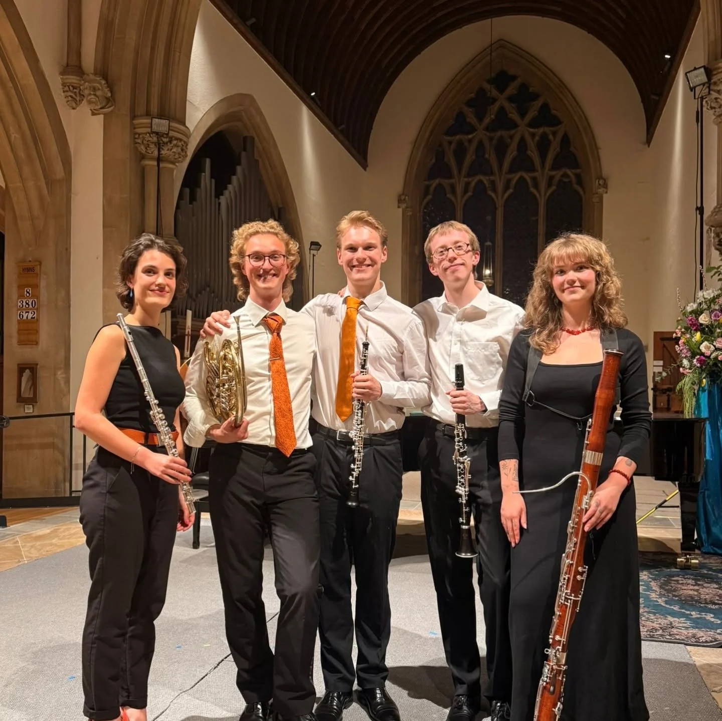 Group of five musicians holding wind instruments in a church or concert hall with wooden arches and stained glass windows.