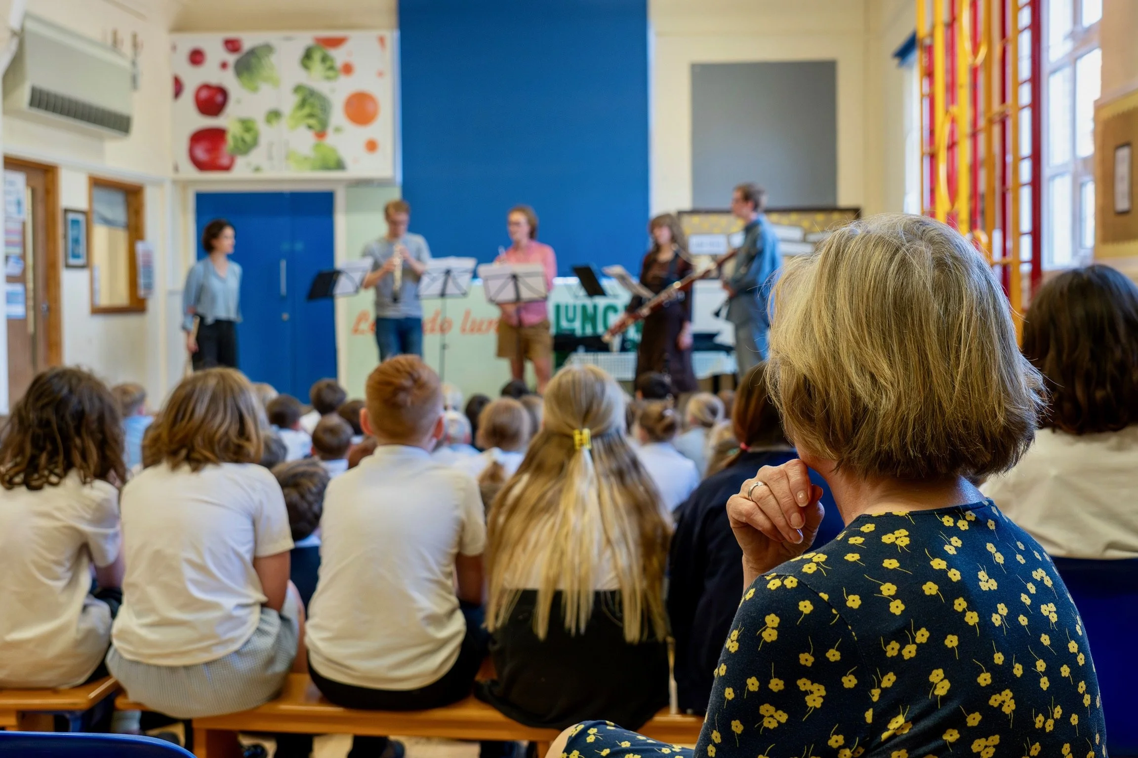 Children sitting on benches watching a live musical performance by a small group of musicians in a school or community hall.