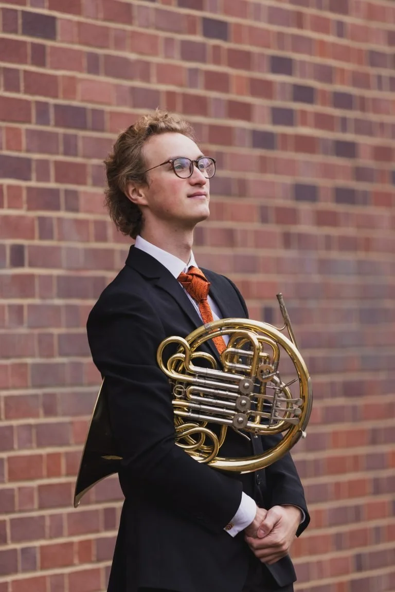 A young man with glasses, wearing a black suit, white shirt, and orange tie, stands outdoors against a brick wall while holding a French horn.