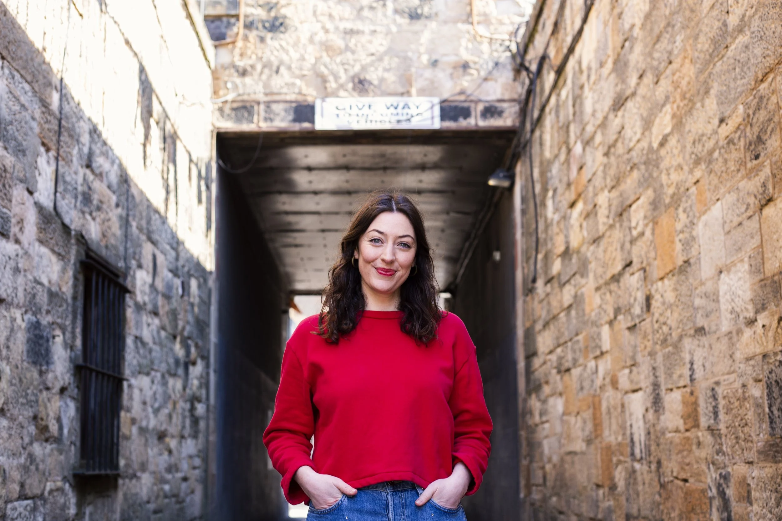 A woman with dark hair wearing a red shirt standing in an alleyway between brick walls.