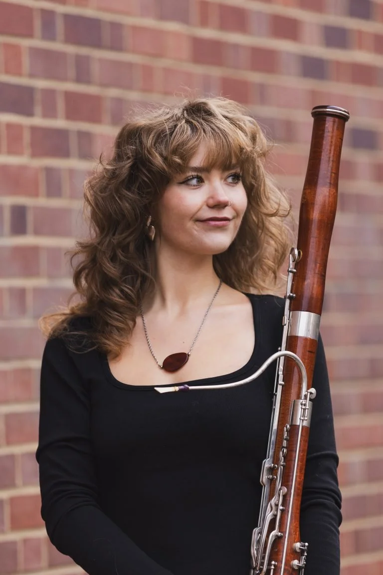 A woman holding a bassoon, standing against a brick wall.