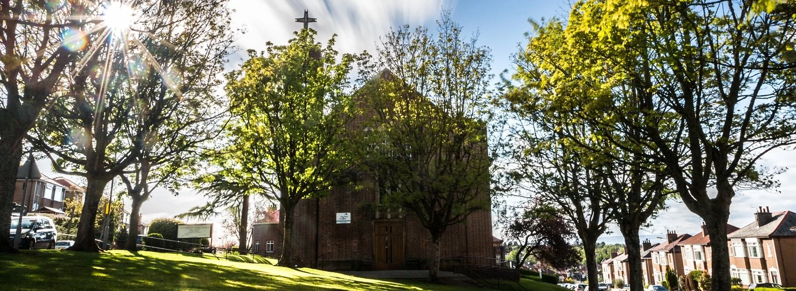 Sunlight shining through trees in front of a brick church with a cross on top, in a residential neighborhood with parked cars.