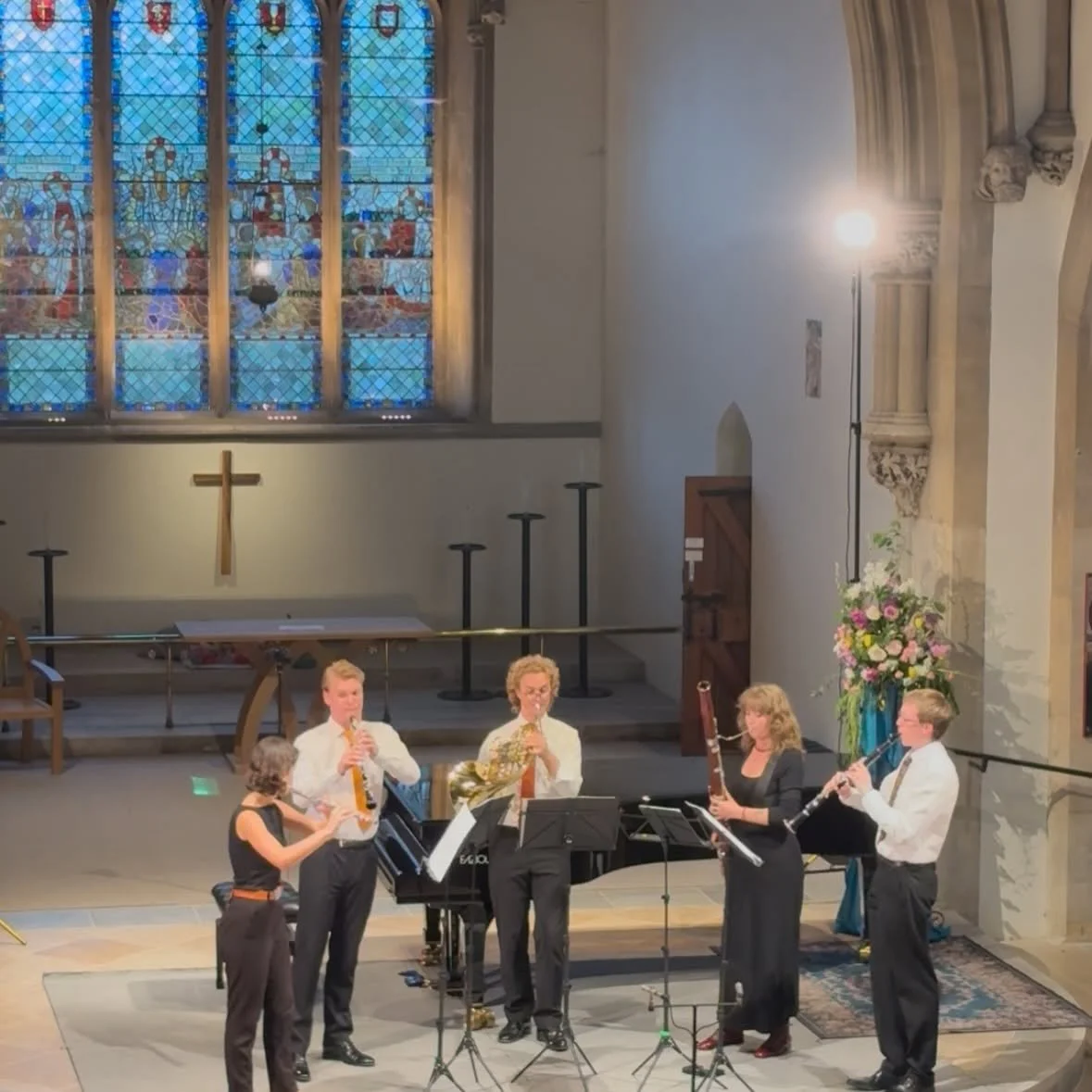 A group of five musicians performing in a church, playing wind instruments including clarinet, saxophone, and flute, with a stained glass window and a wooden cross in the background.