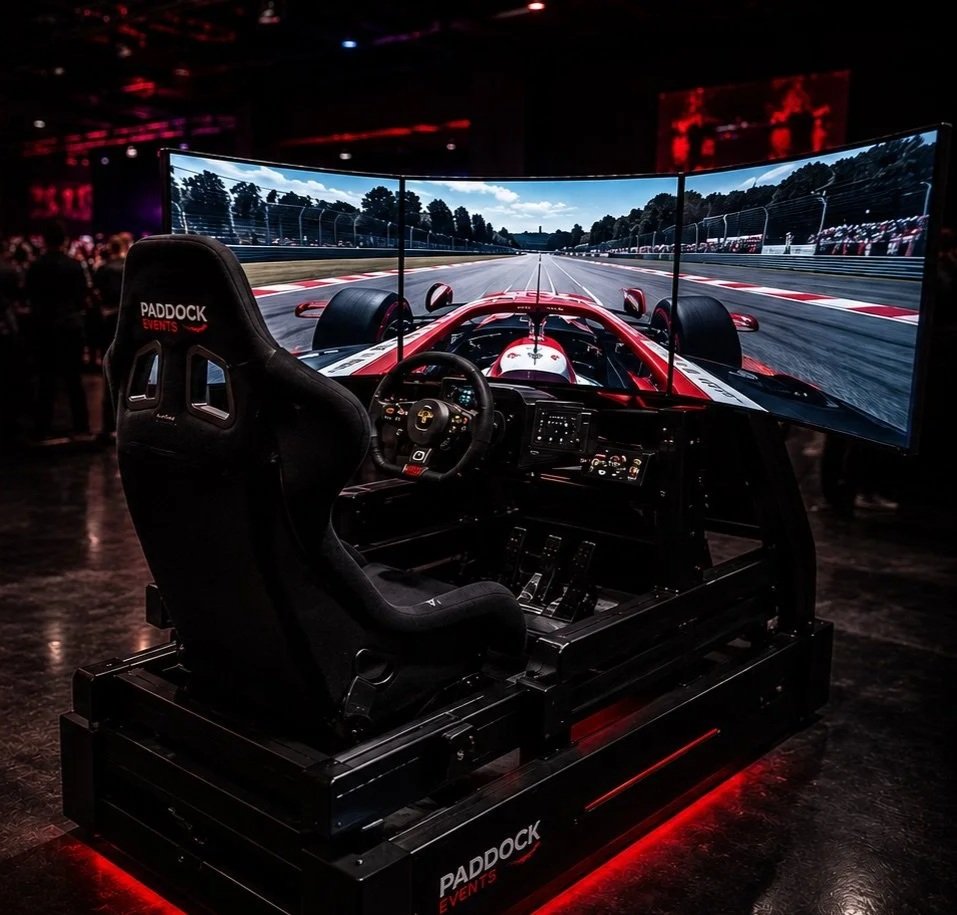 A racing simulator with three large curved screens displaying a race track, a racing seat labeled 'Paddock Events,' and a steering wheel and controls, set inside a dimly lit room.