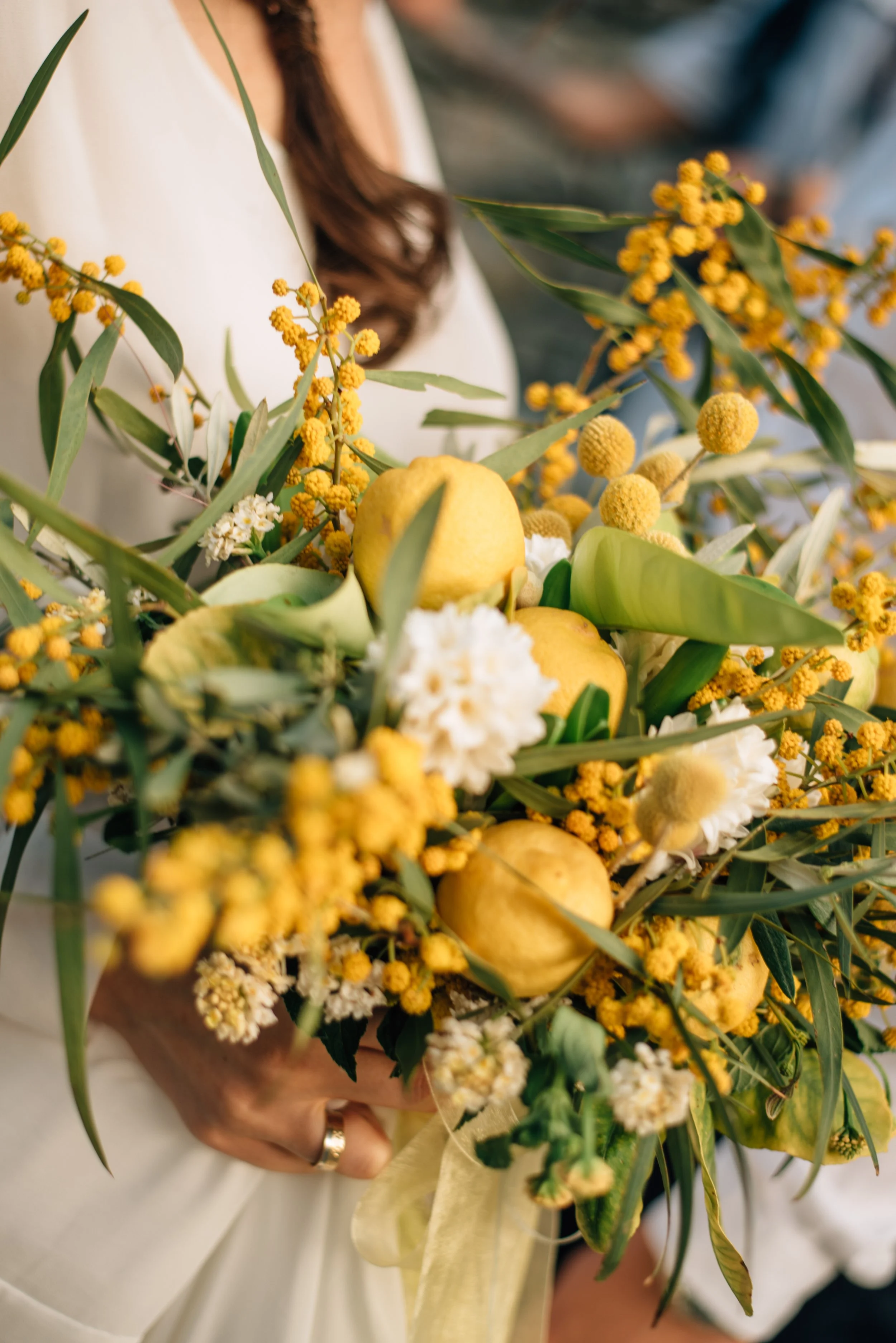 Eine Person hält einen Blumenstrauß mit gelben Blumen, grünem Laub und weißen Blüten, außerdem enthält der Strauß mehrere Zitronen. Die Person trägt ein weißes Kleid und hat braune, gewellte Haare.