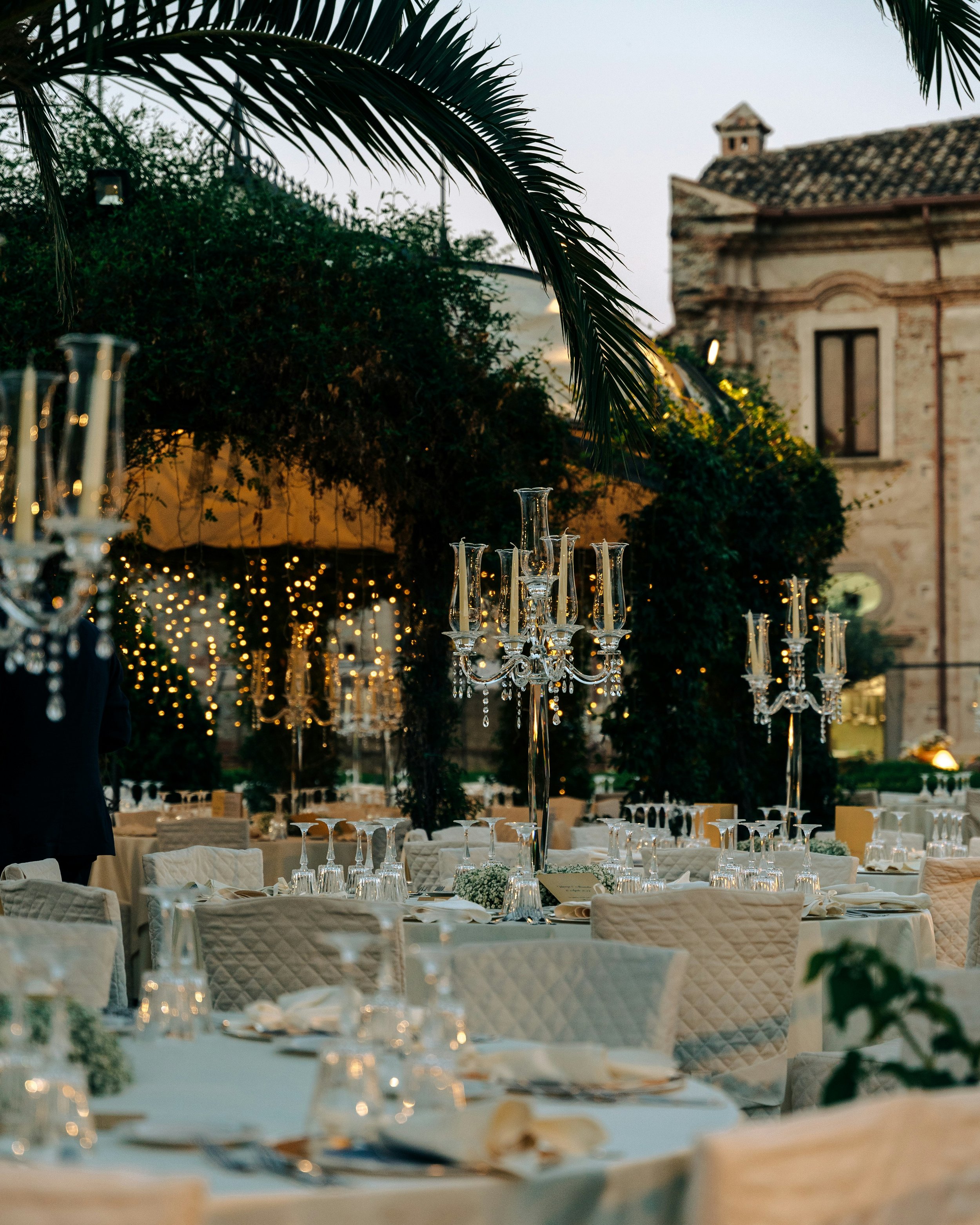 Eleganter festlicher Tisch mit Kristallkerzenleuchtern, weißen Tafeldecken und Dekorationen in einem Garten mit Palmblättern und historischen Gebäuden im Hintergrund bei Dämmerung.