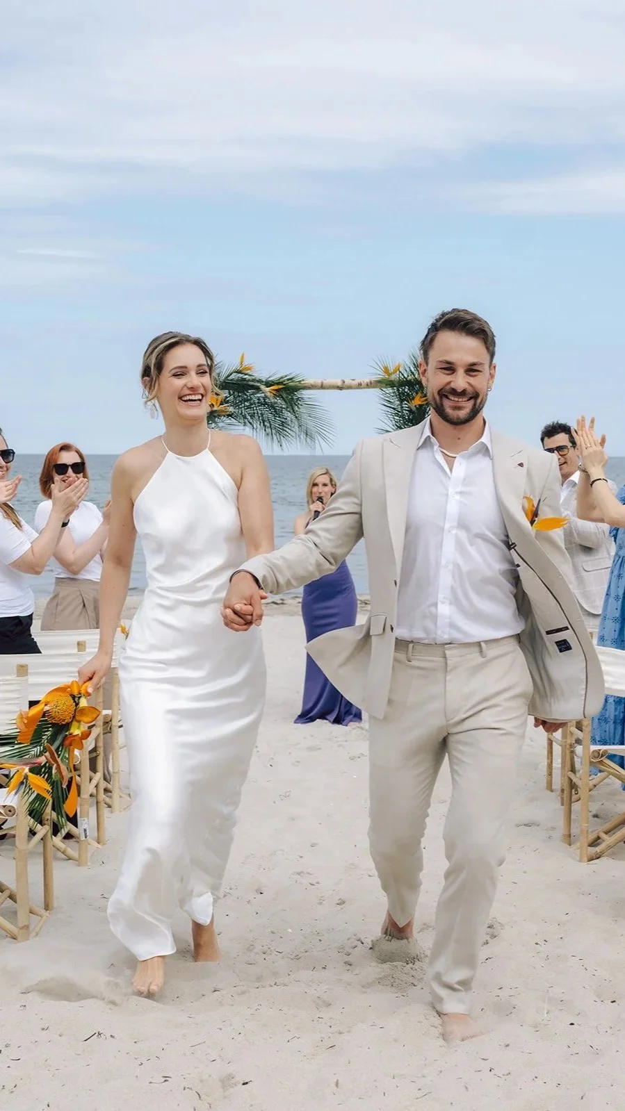 Ein glückliches Paar bei einer Strand-Hochzeit, umgeben von lachenden Gästen, die bei Sonnenbrillen und sommerlicher Kleidung am Strand feiern.