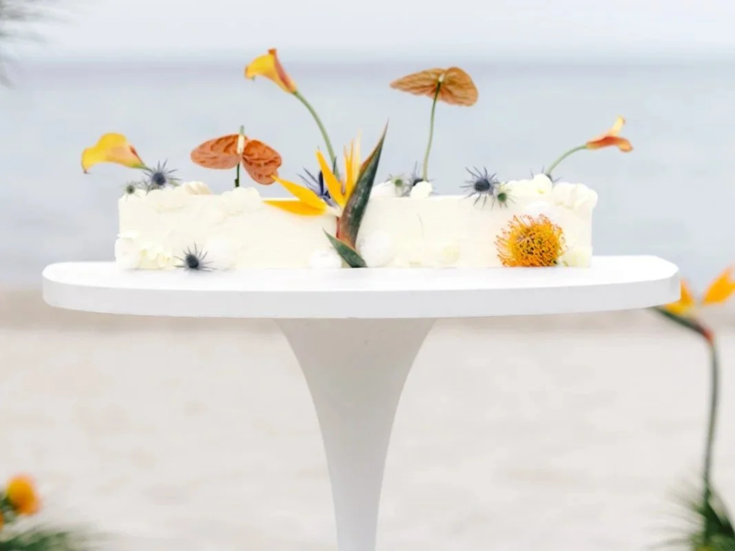 White cake decorated with edible flowers and small black and yellow flowers on a white cake stand.
