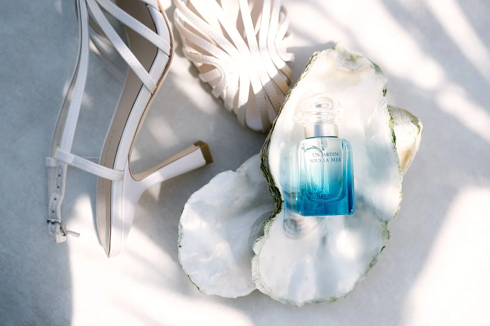 A bottle of Hermès Un Jardin Sous La Mer perfume stands inside a large seashell with white and green edges. Nearby are white high-heeled sandals and a small white tube, all arranged on a light surface with dappled sunlight creating shadows.