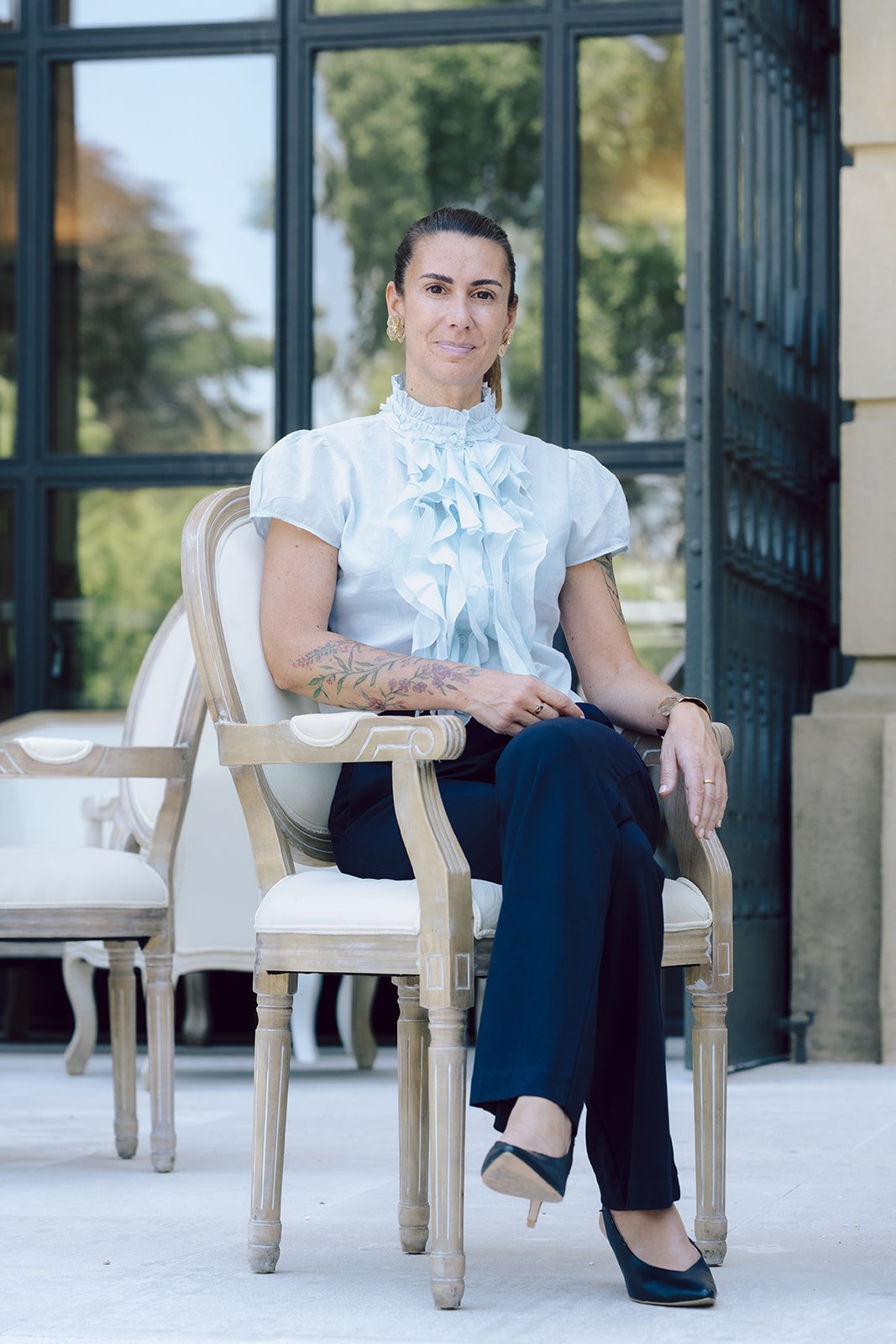 Frau mit Tattoos, gebügeltem weißen Blüs, schwarzen Hosen und High Heels sitzt auf einem antiken Stuhl im Freien bei Sonnenlicht.