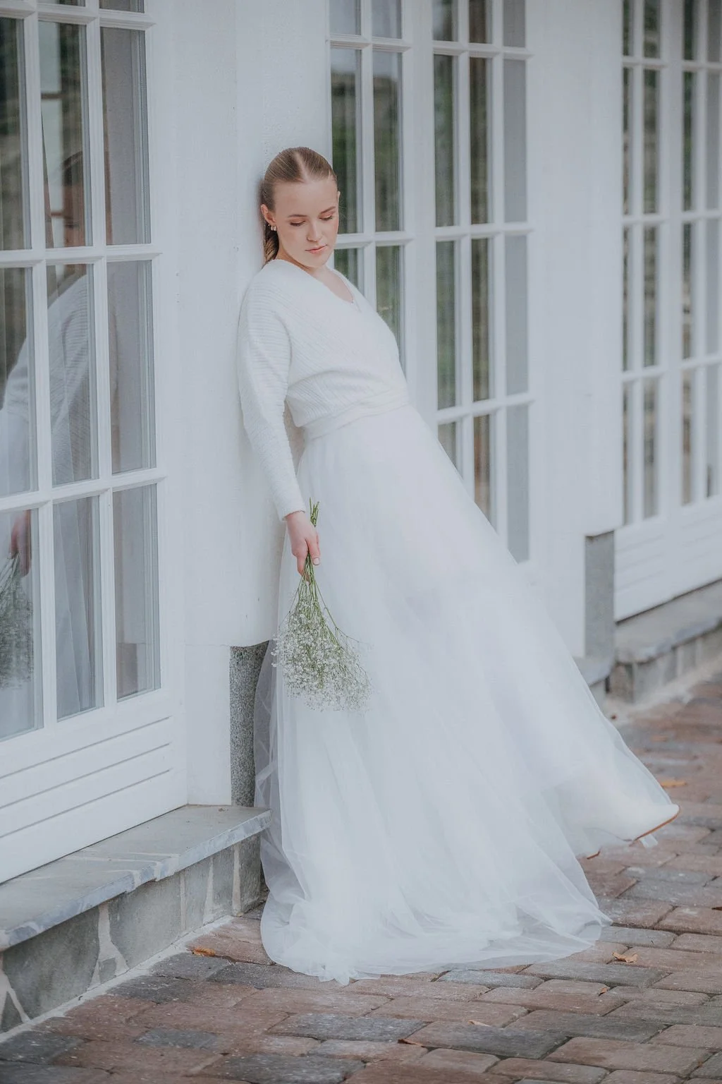 Eine junge Frau in weißem Hochzeitskleid lehnt an einer weißen Wand vor großen Fenstern und hält einen kleinen Blumenstrauß in der Hand.
