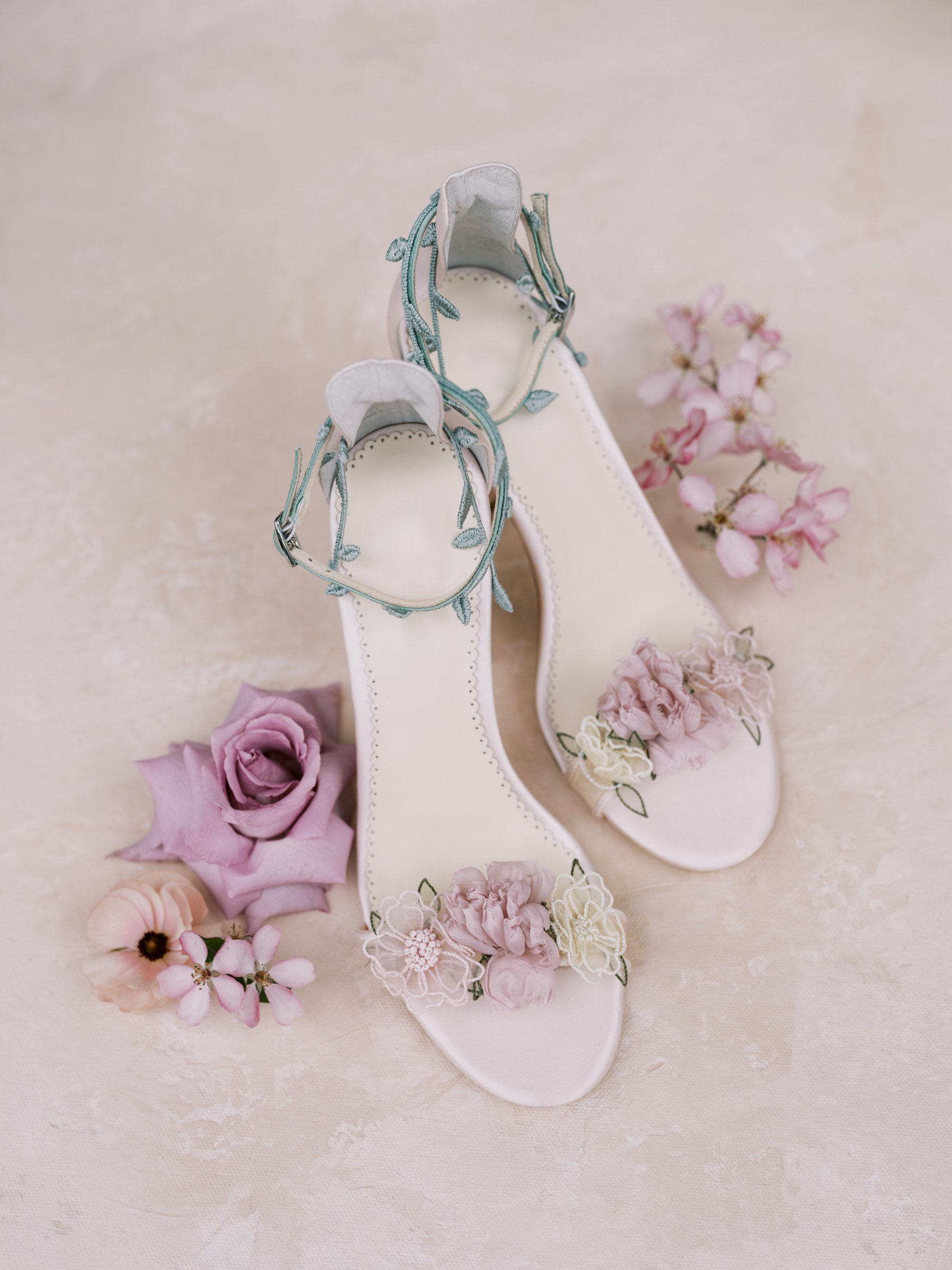 Weiße High Heels mit blauen Blumenverzierungen, umgeben von rosa und weißen Blumen auf hellem Hintergrund.
