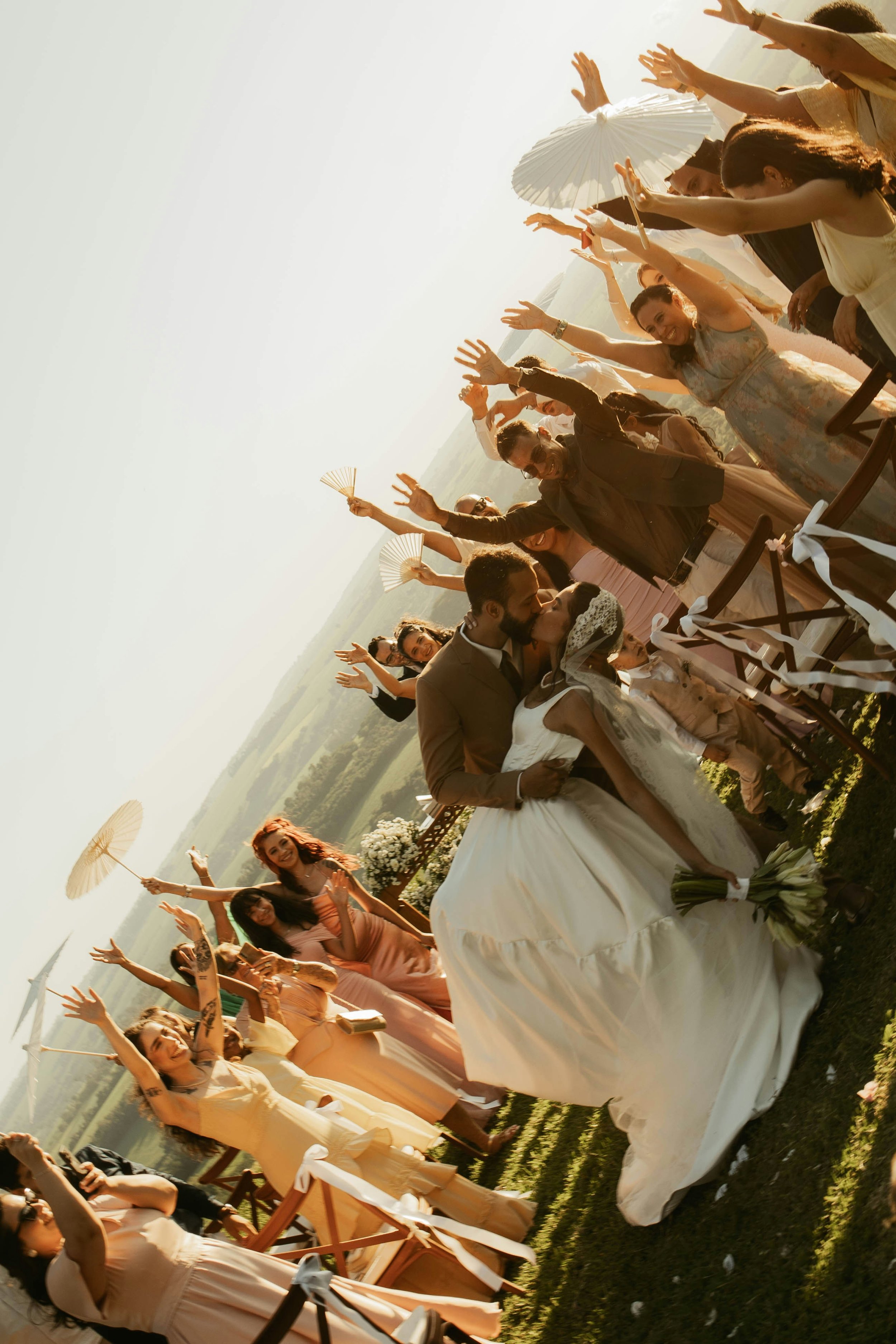 Hochzeitspaar küsst sich vor einer Gruppe lachender Gäste, die mit Sonnenschirmen feiern, im Freien bei Sonnenuntergang.
