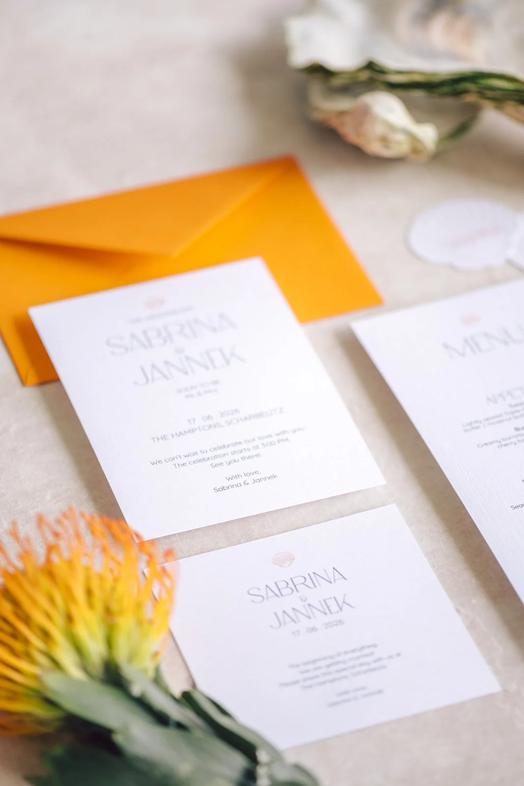 Ein Einladungsticket für eine Hochzeit mit dem Namen Sabrina und Jannek, Datum 17.06.2023, mit Blumen und einem orangefarbenen Umschlag im Hintergrund.