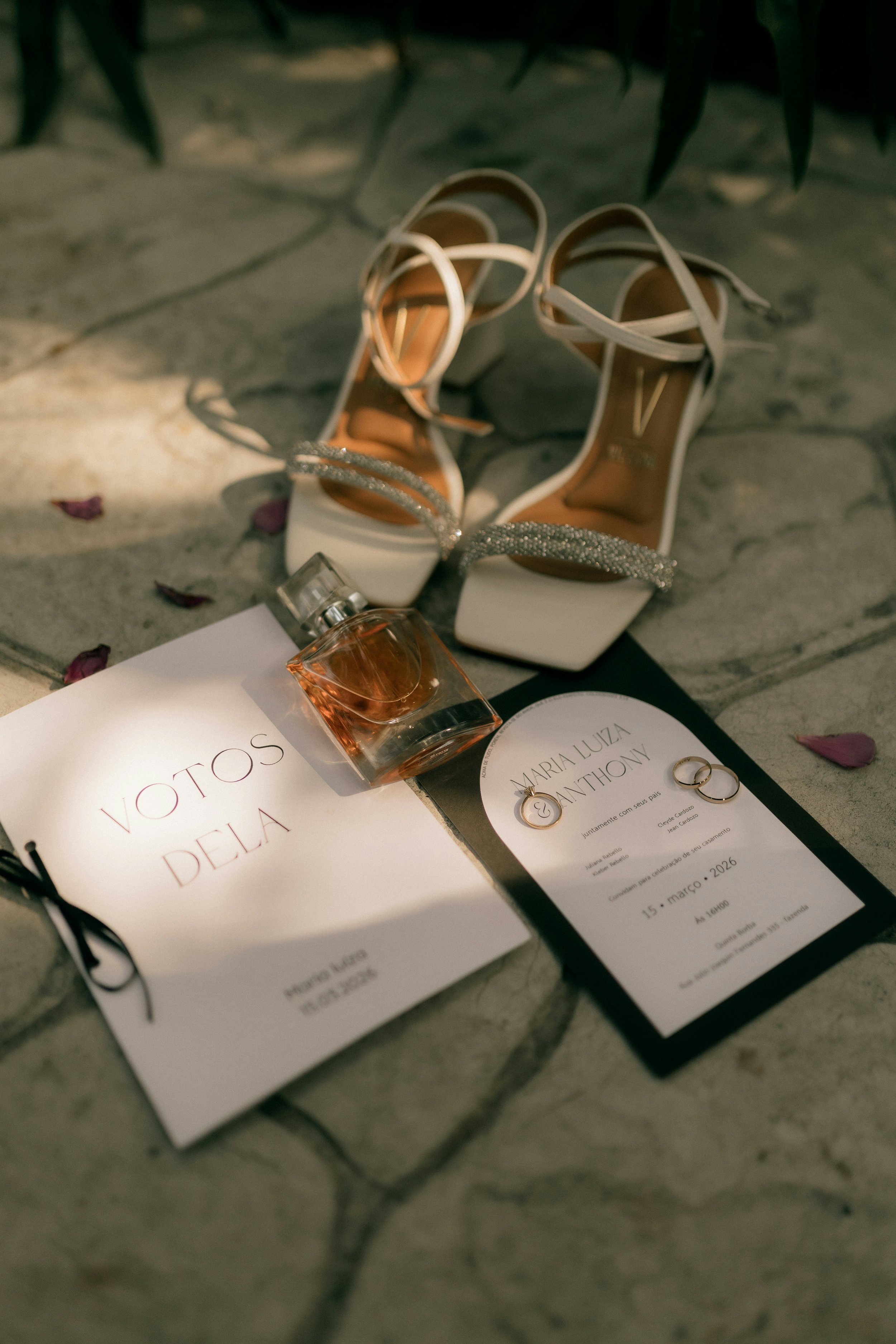 Ein Paar weiße High Heels mit silberner Schnalle, ein Parfümfläschchen und eine Hochzeitsurkunde auf einem Steinboden, umgeben von Blumenblättern.