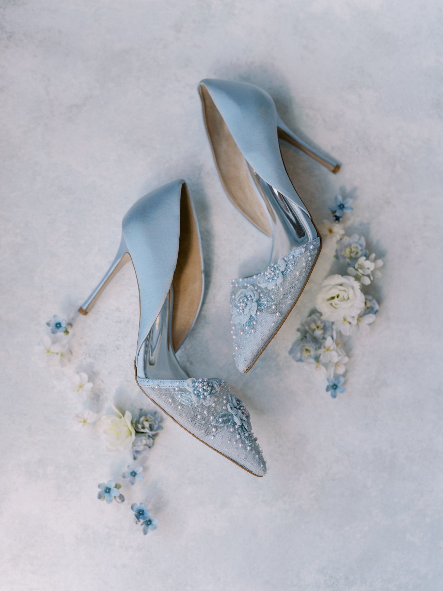 Ein Paar elegante, hellblaue High Heels mit Schmuckdetails auf einer weißen Oberfläche, umgeben von kleinen weißen und blauen Blumen.