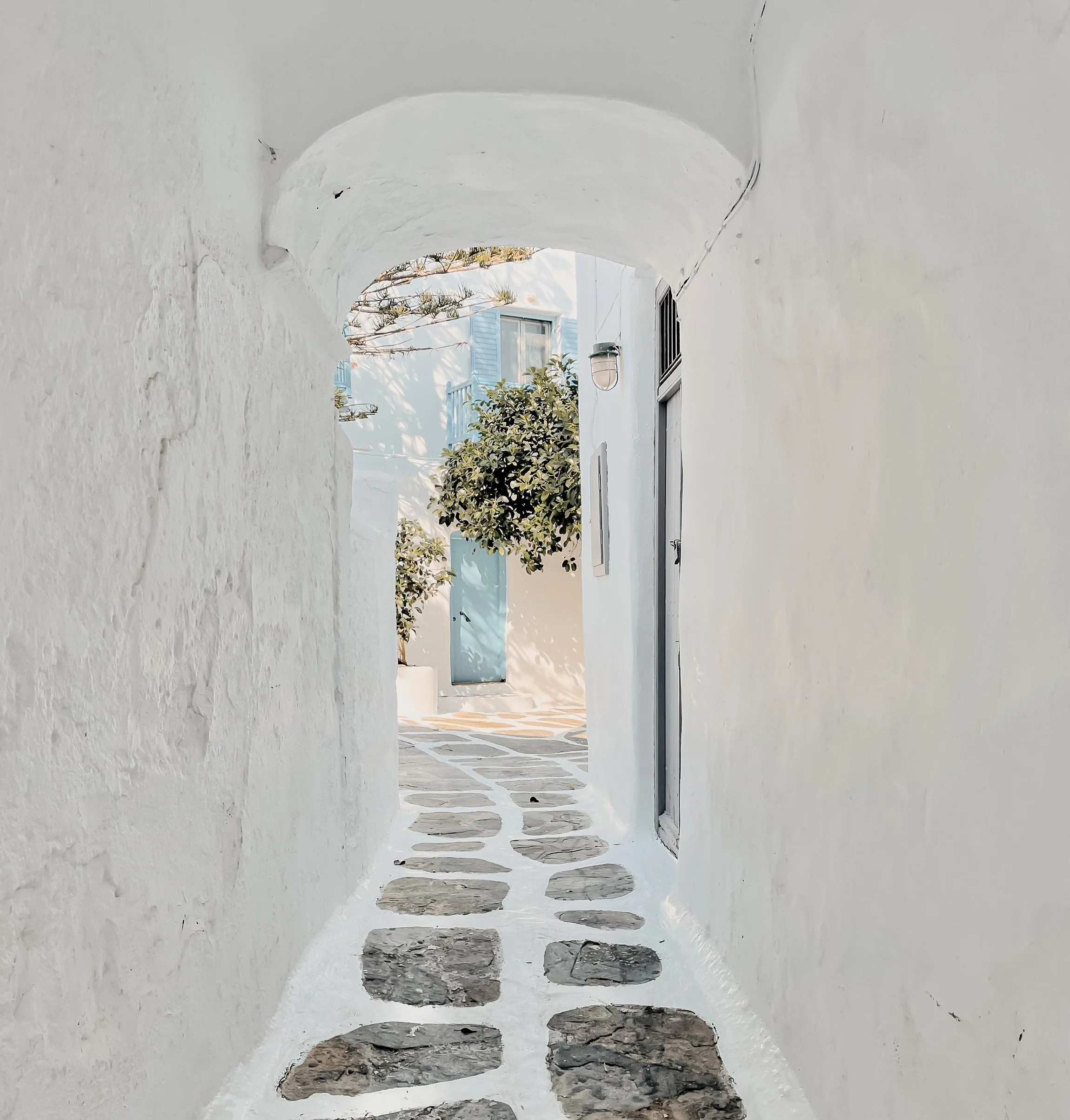 A whitewashed archway with a view of a sunny courtyard, featuring a white building with blue shutters, a hanging light fixture, and greenery.