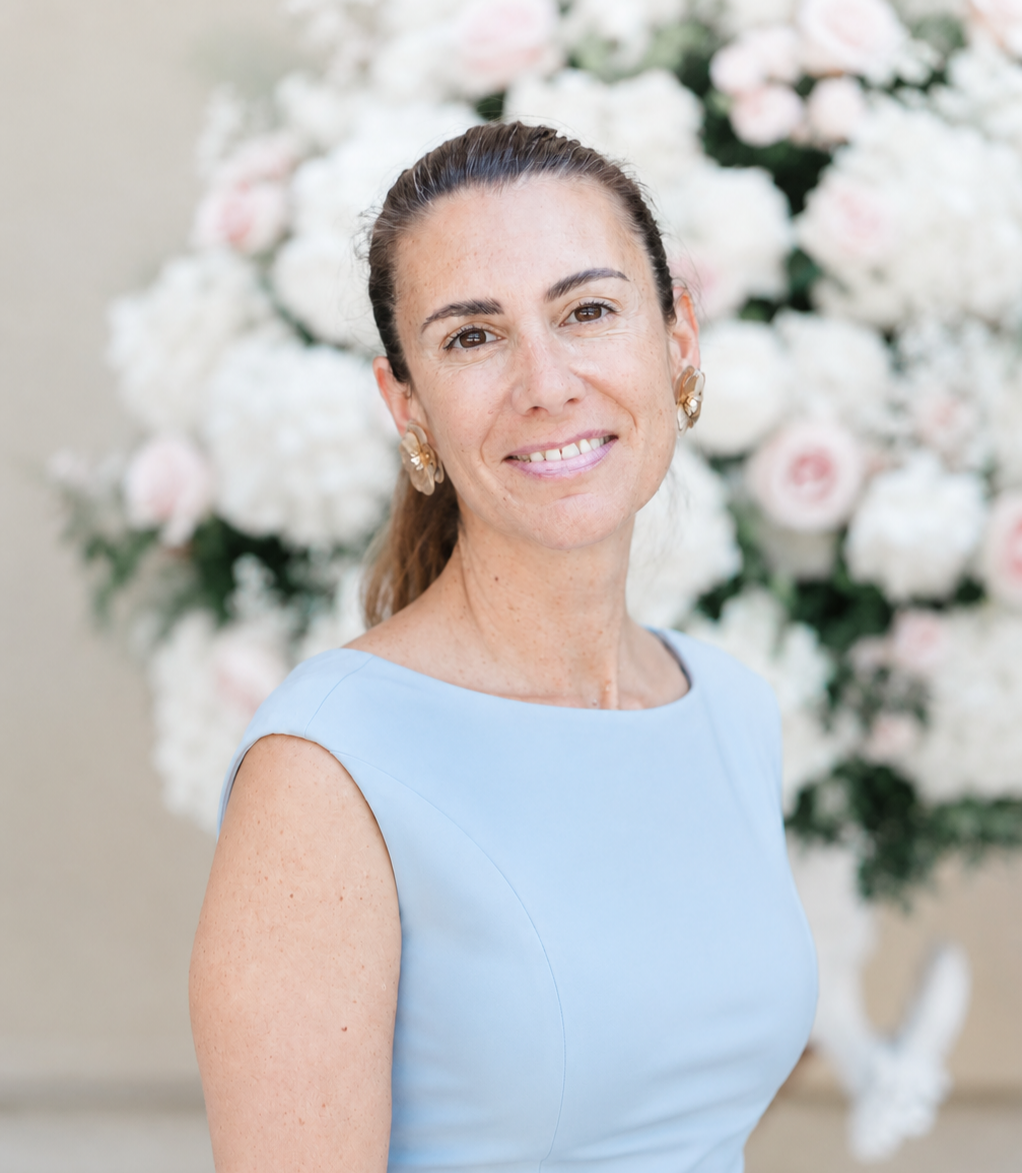 A woman with brown hair pulled back, wearing a light blue sleeveless dress and floral earrings, smiling at the camera, with a background of white and pink flowers.