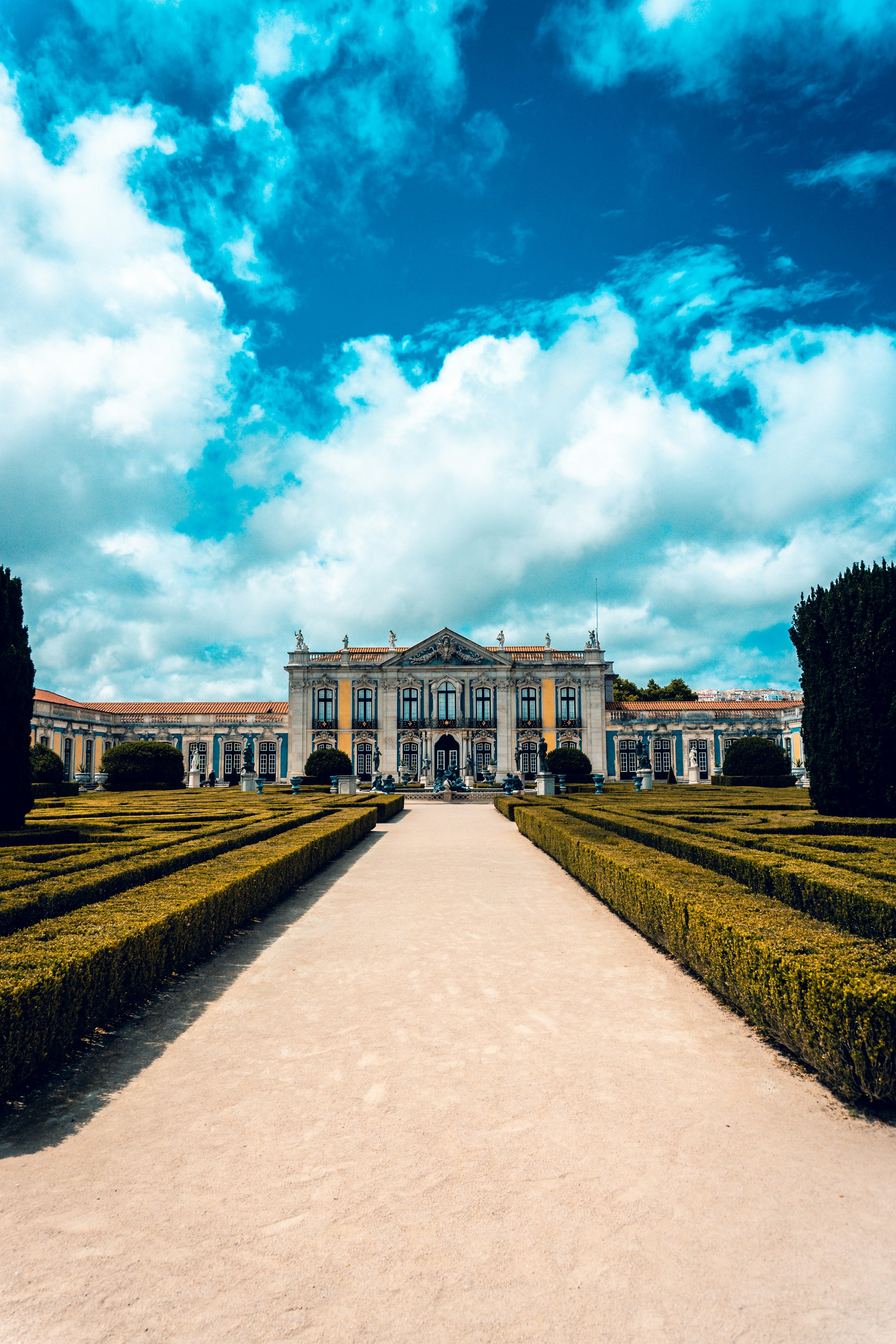 Ein barockes Gebäude mit goldverzierten Details, umgeben von gepflegten Gärten und grünen Bäumen, unter einem bewölkten blauen Himmel.