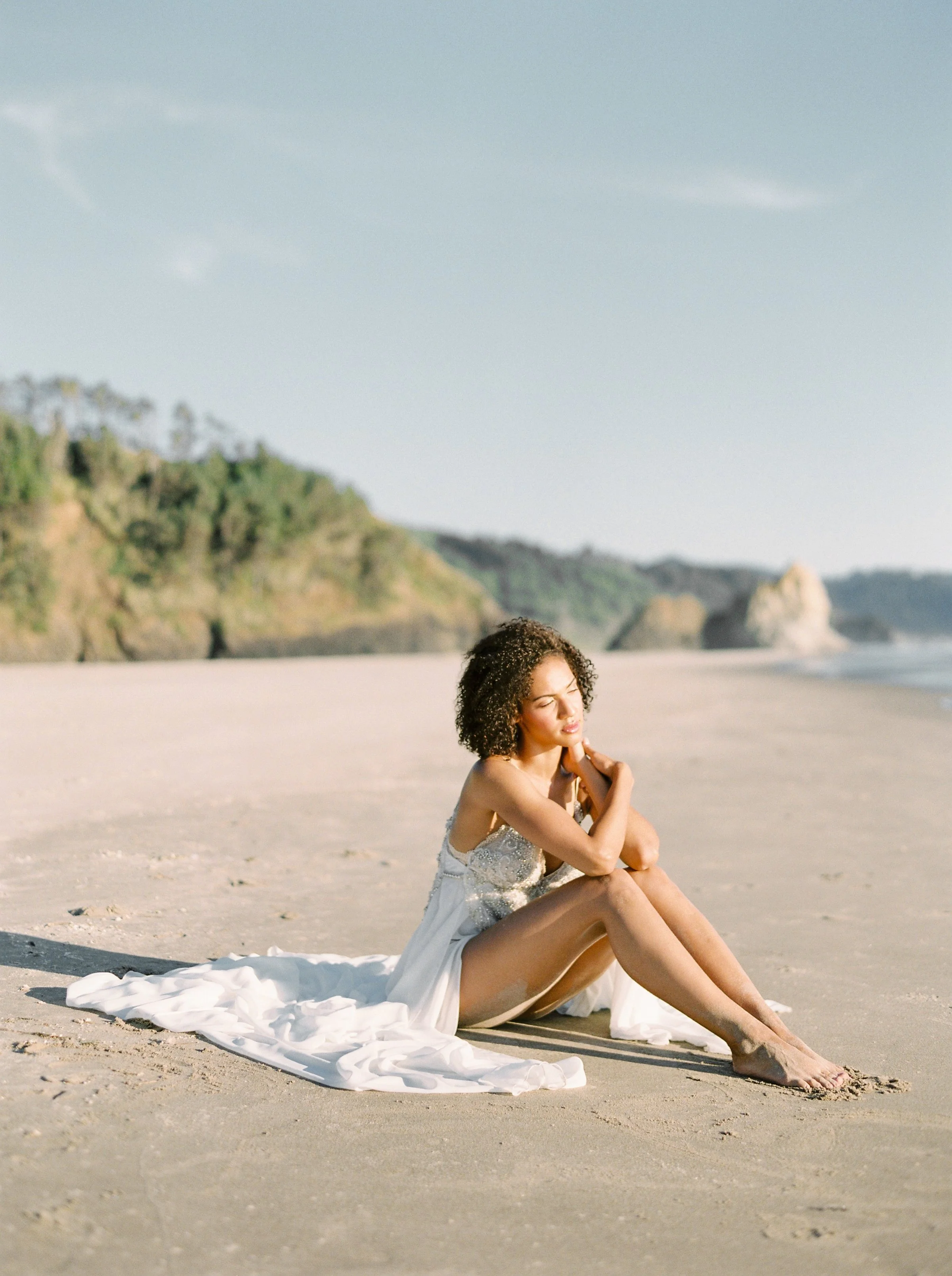 Eine junge Frau sitzt entspannt am Strand auf einem weißen Tuch, trägt ein weißes Kleid und hat lockiges Haar. Im Hintergrund sind Klippen und das Meer sichtbar, die Stimmung ist friedlich und sonnig.