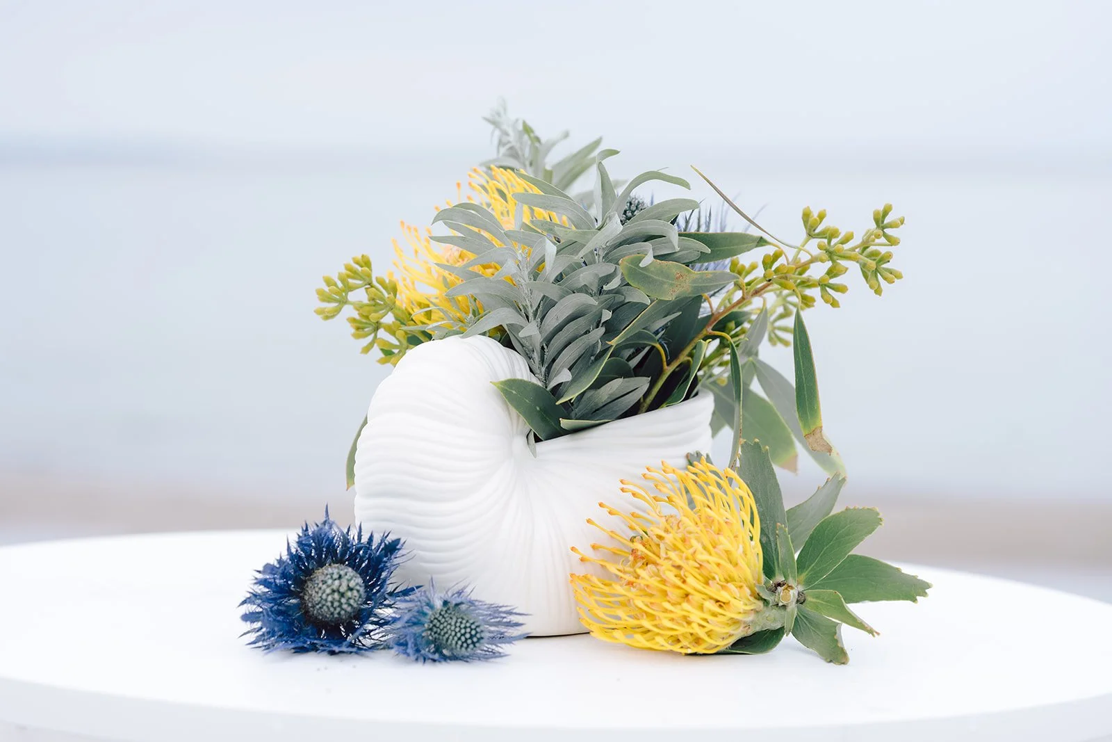 Arrangement of various flowers and greenery in a white ceramic vase on a white surface