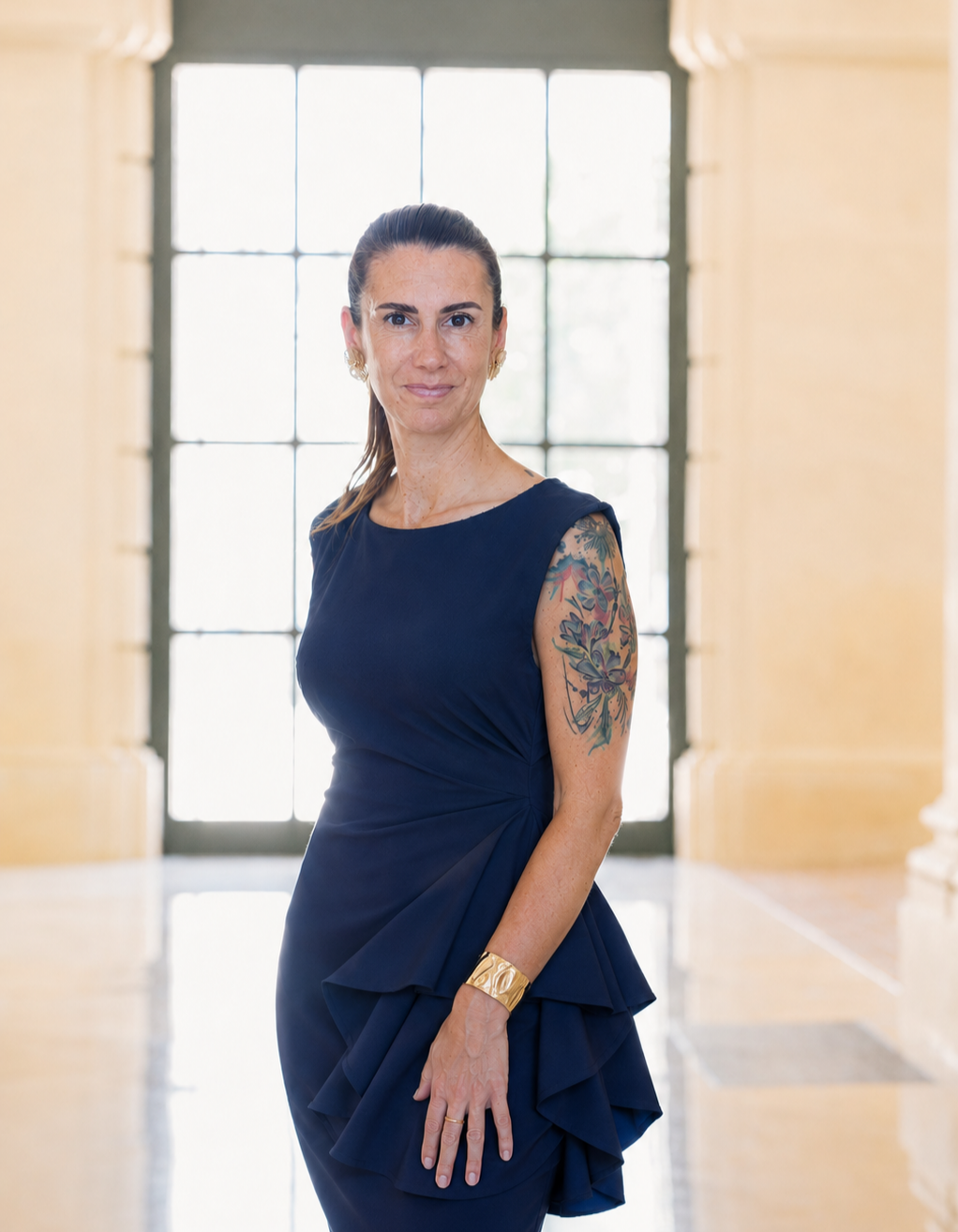 Frau in einem eleganten, dunkelblauen Kleid mit Tattoo auf dem Arm, stehend vor einem großen Fenster, im hellen Raum.