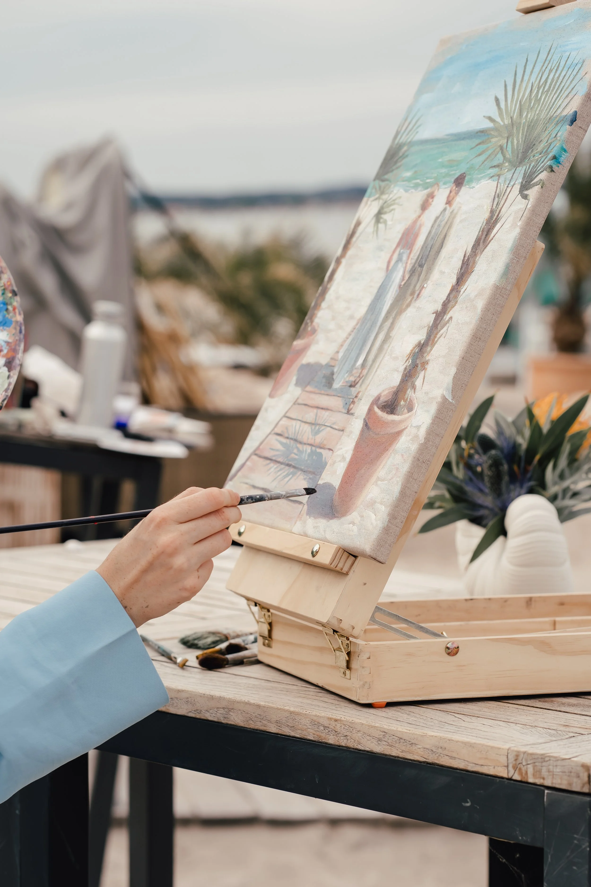 Eine Person malt eine Strandlandschaft mit Palme und Meer auf eine Leinwand, die auf einem kleinen Holzrahmen steht. Der Hintergrund zeigt eine Aussicht auf den Strand bei bewölktem Himmel.