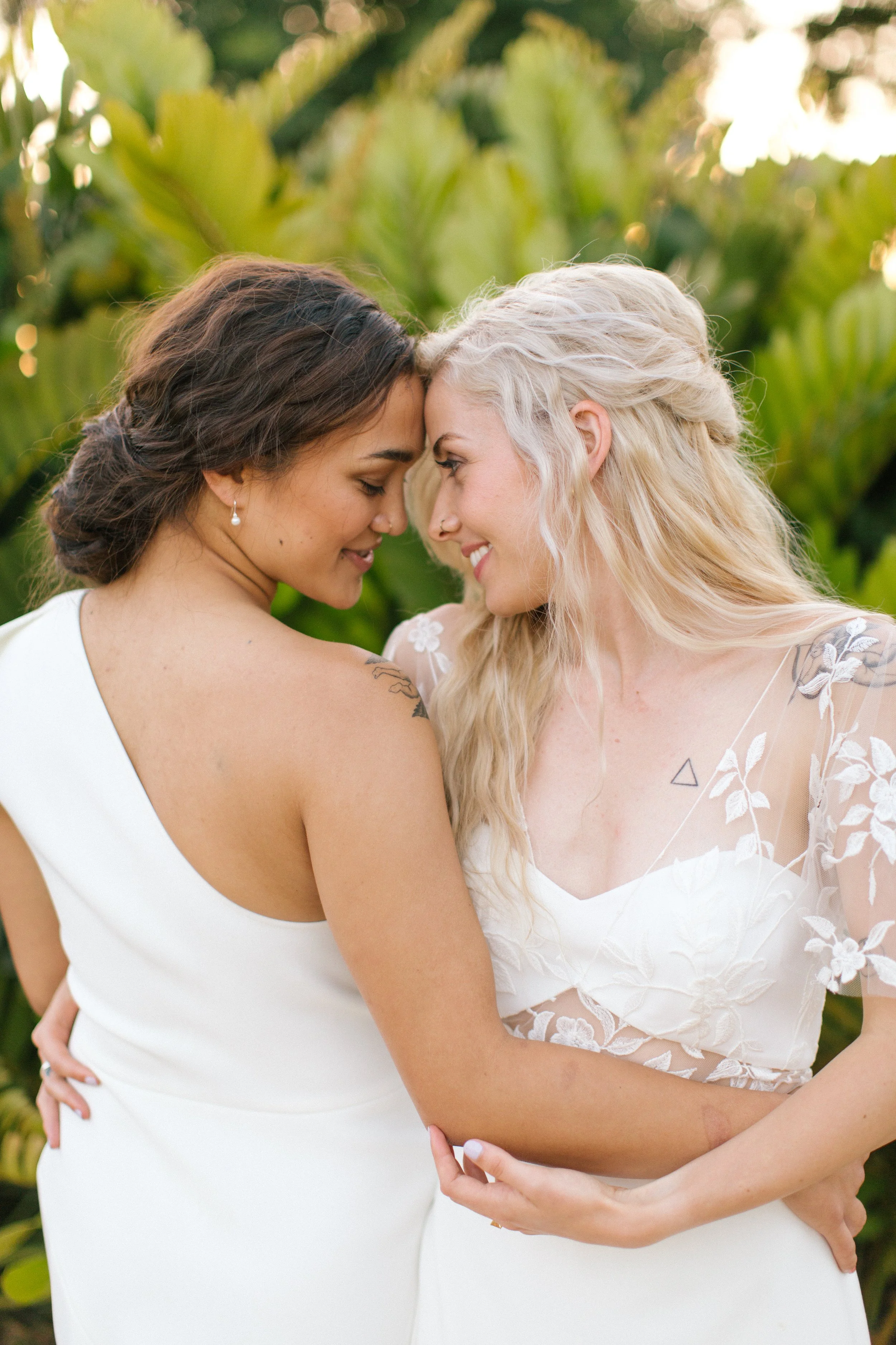 Zwei Frauen stehen eng beieinander mit Stirn an Stirn, lächeln und umarmen sich vor grünem Blätterhintergrund. Eine trägt ein schlichtes, weißes Kleid, die andere ein elegantes Kleid mit Blumenmuster.