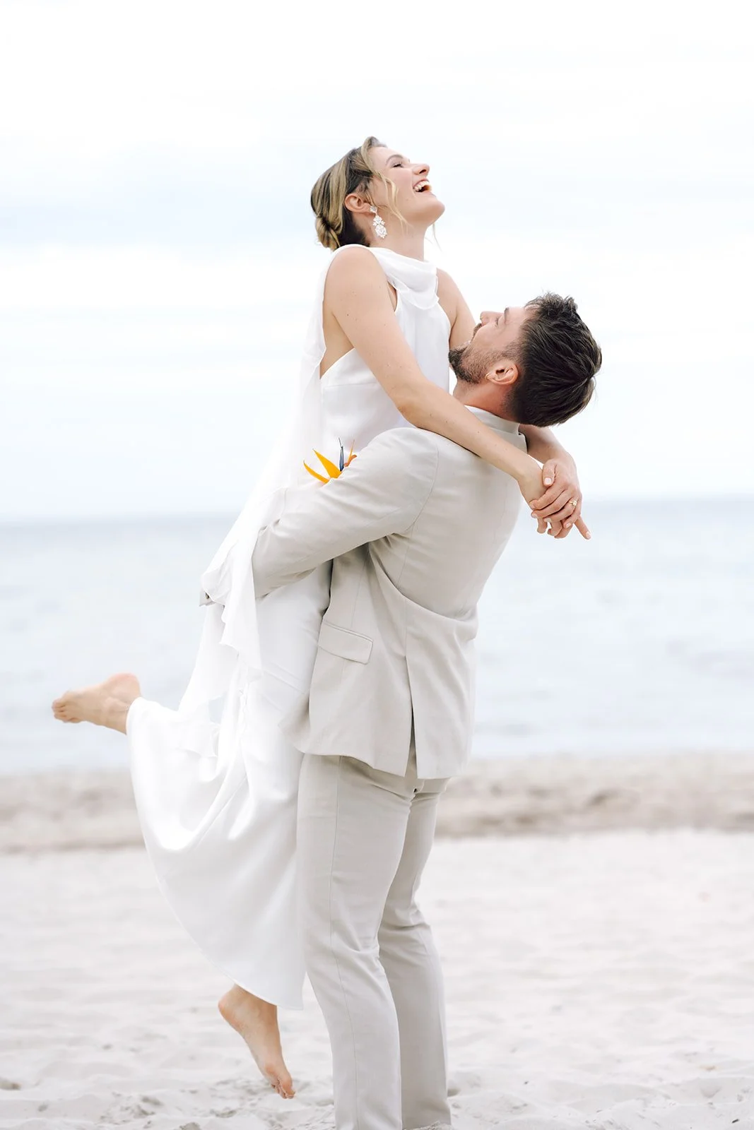 Ein lachendes Paar am Strand, der Mann trägt einen hellen Anzug, die Frau ein weißes Kleid, die Frau wird vom Mann getragen.