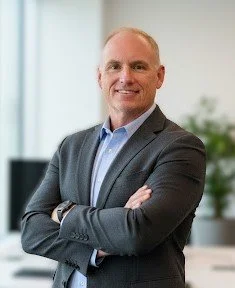 A professional man in a suit standing with arms crossed in an office setting.