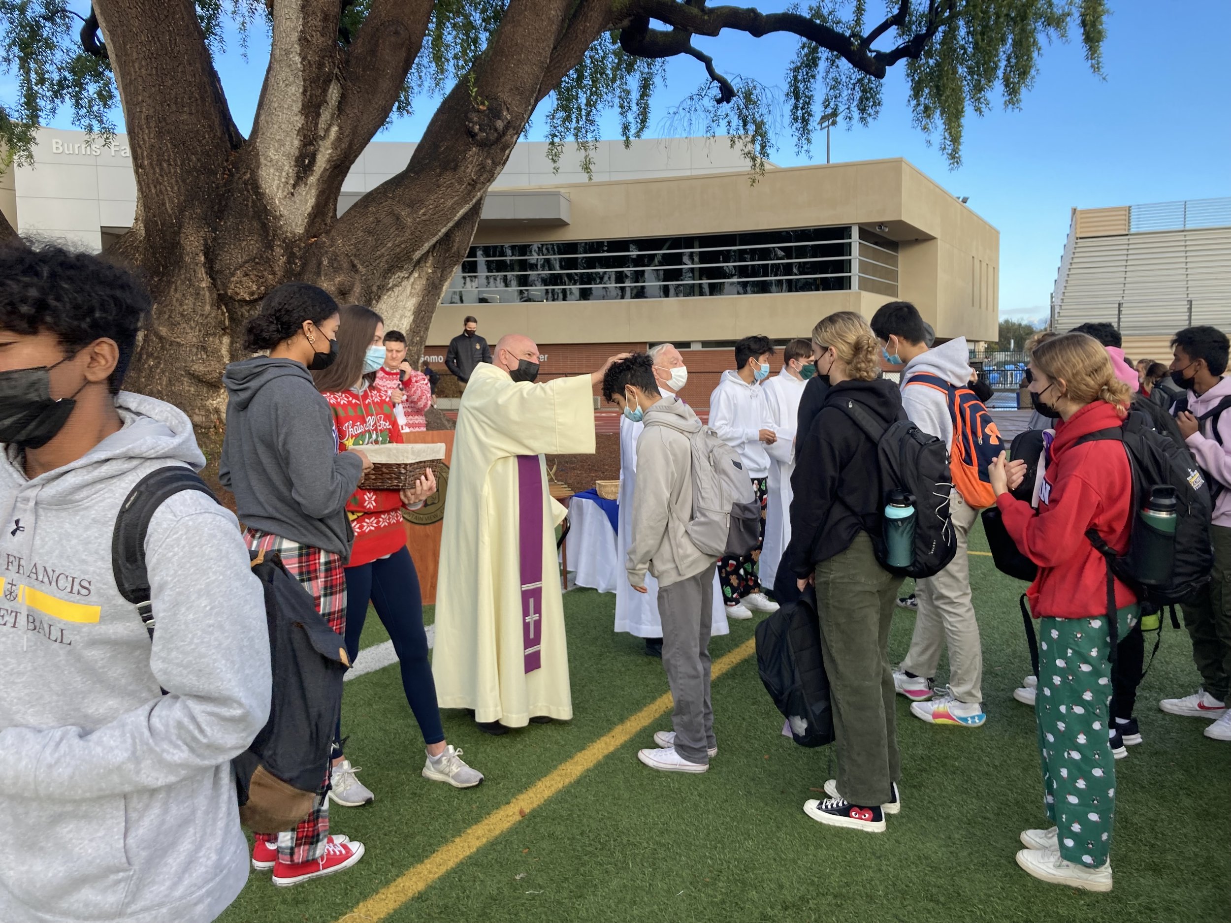 A group of students standing in line outdoors, wearing masks, with a religious figure, possibly a priest, blessing them. The scene takes place on a school track field next to a large tree and modern school building.