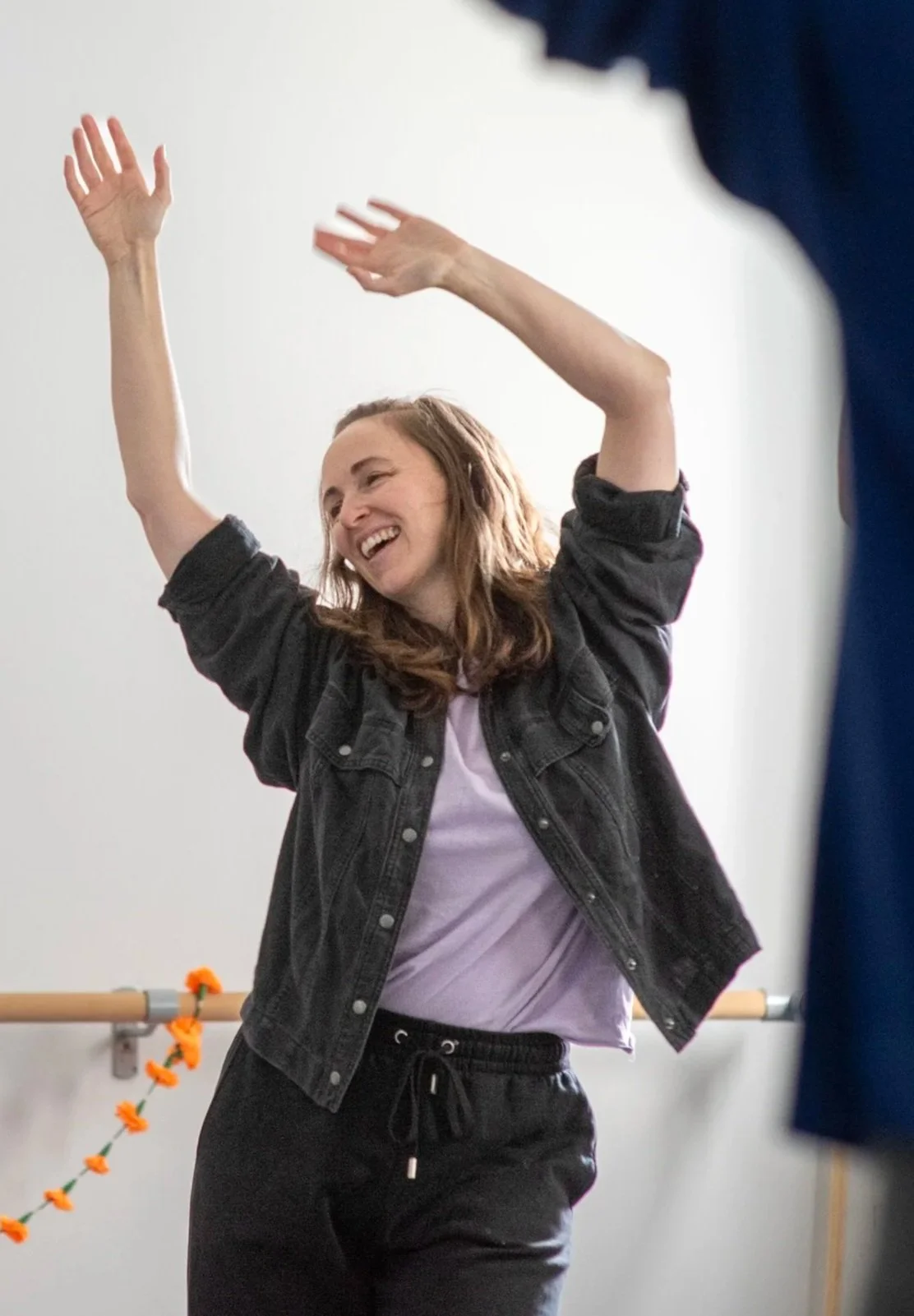 Sarah, with brown hair, loose to her shoulders, is wearing a black jacket and black tracksuit bottoms, smiling and raising her arms in a joyful pose indoors. There's also side of someone's body in the foreground, also arms raised.