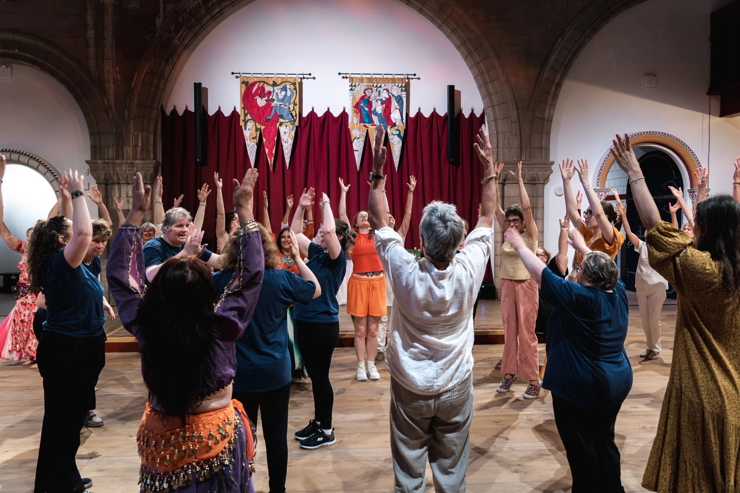 Group of people participating in a dance or activity inside a hall with stage, red curtains, and medieval-style banners hanging on the wall.