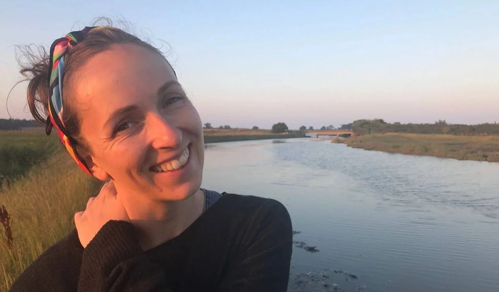 Sarah, a woman with pale skin, brown hair tied back with a colourful headscarf is standing near a river at sunset.