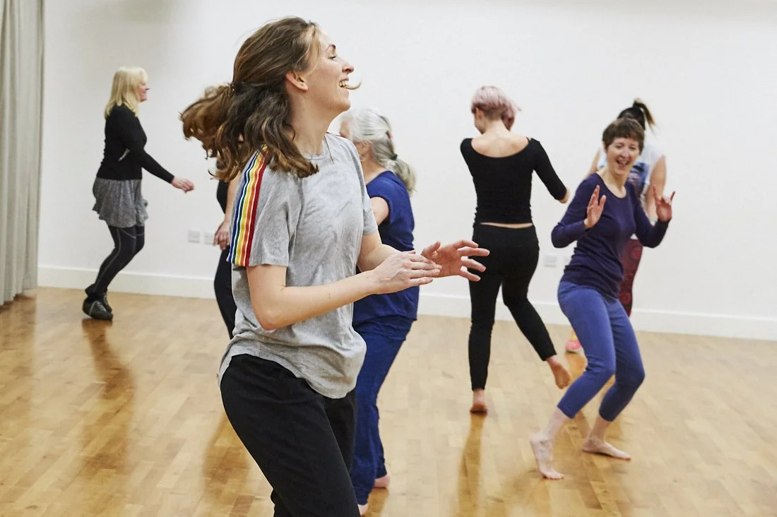 Group of women dancing and laughing in a room with wooden floors and white walls. They're all doing different things but smiling and having fun together.
