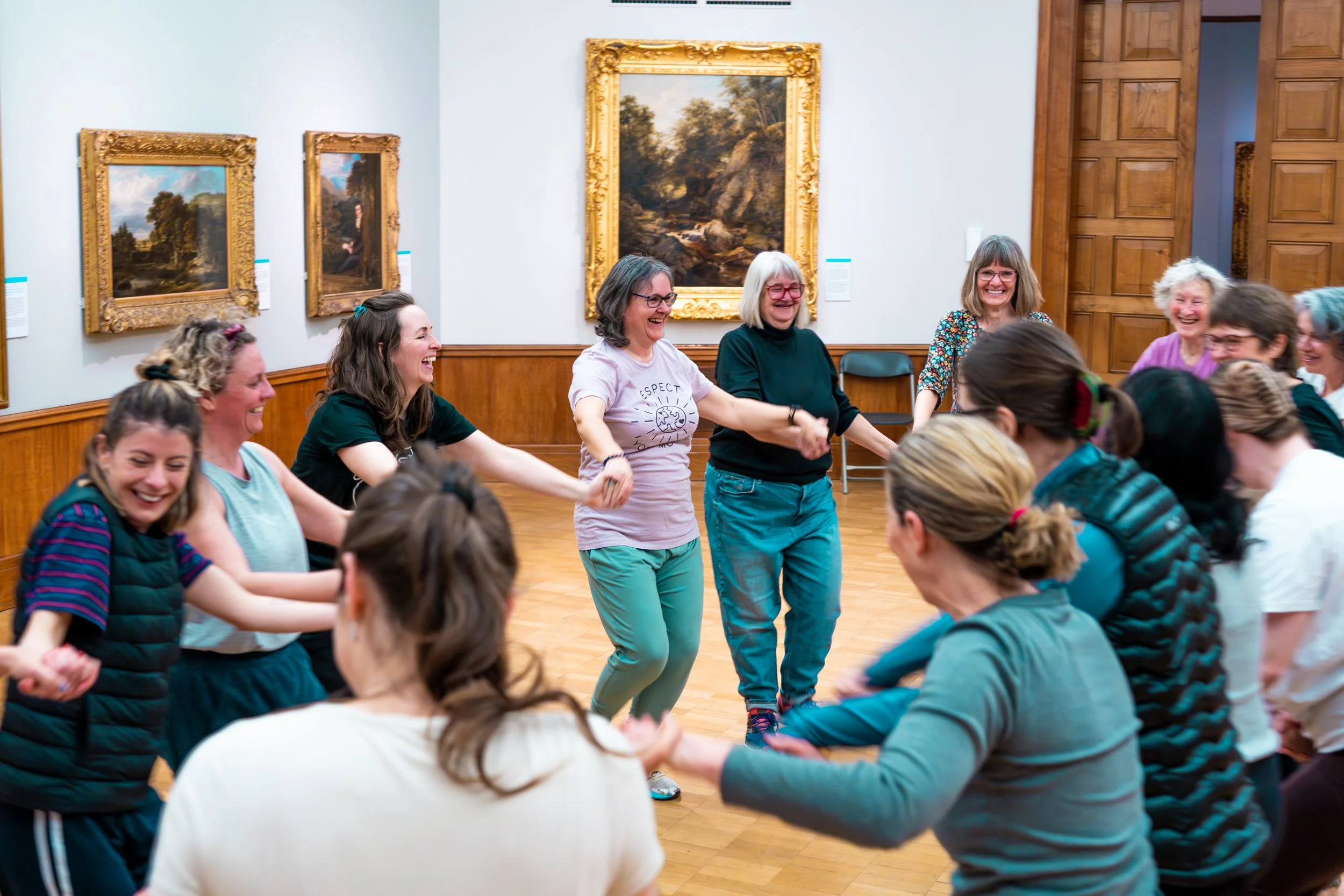 A group of women holding hands and smiling in a circle, participating in a dance or group activity in an art gallery with framed landscape paintings on the walls.
