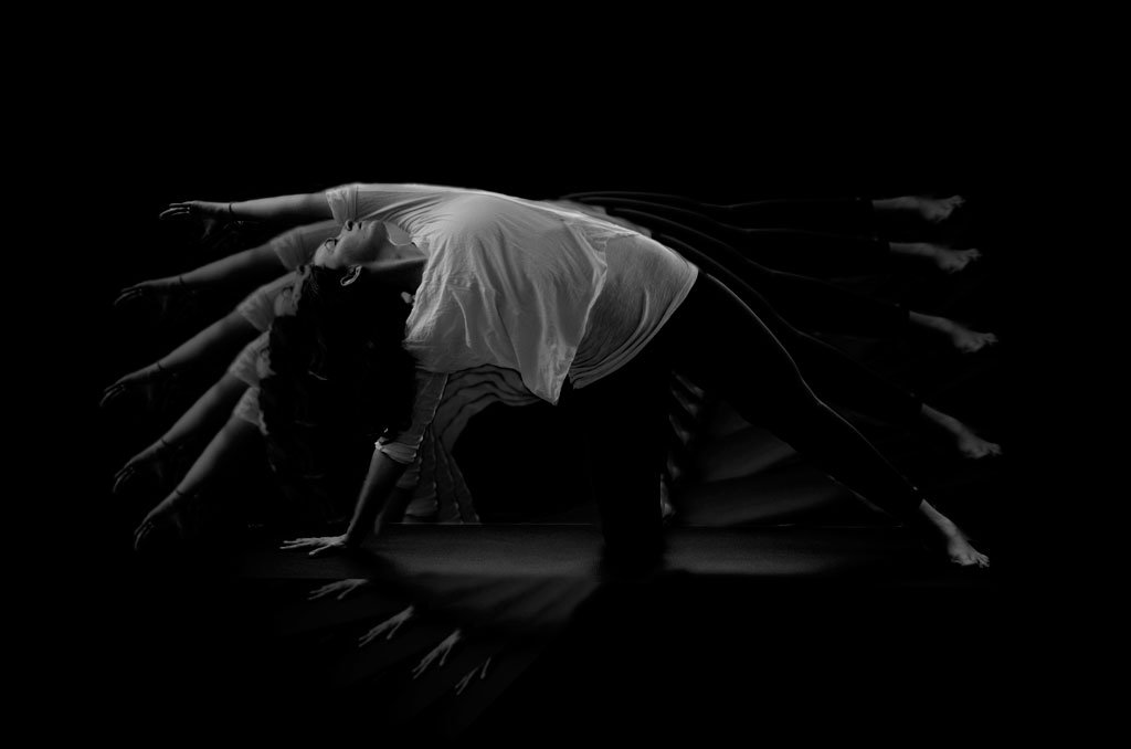 A person in a long, sideways stretch in a dark room with mirrored reflections creating a layered effect, as though they have 5 body's, not just 1